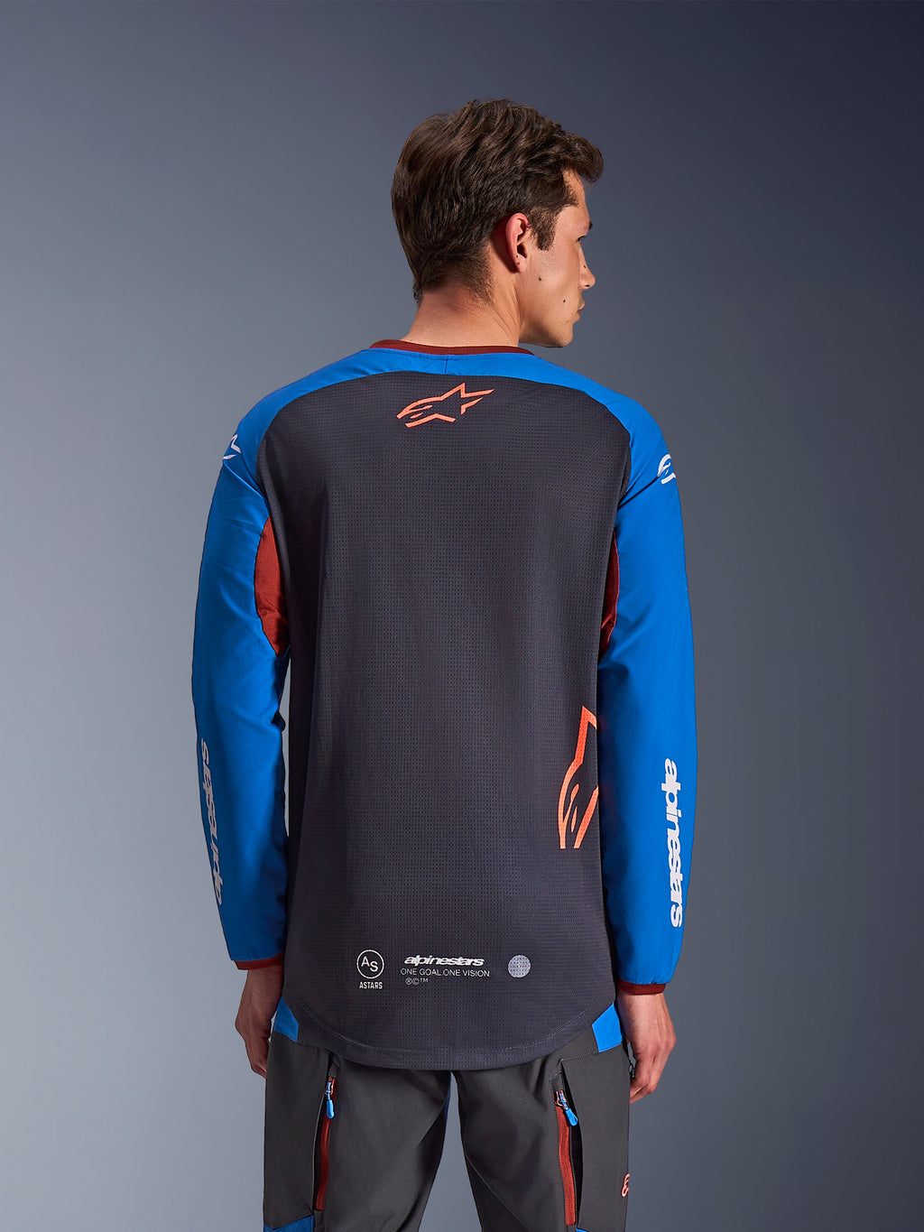 Alpinestars Maxdura Dual Jersey in Deep Gray and Royal Blue, back view of male model, mesh torso construction with orange Astars logo, long sleeves with white branding, maroon collar and cuffs, worn with gray technical pants, dual-sport and adventure motorcycle gear