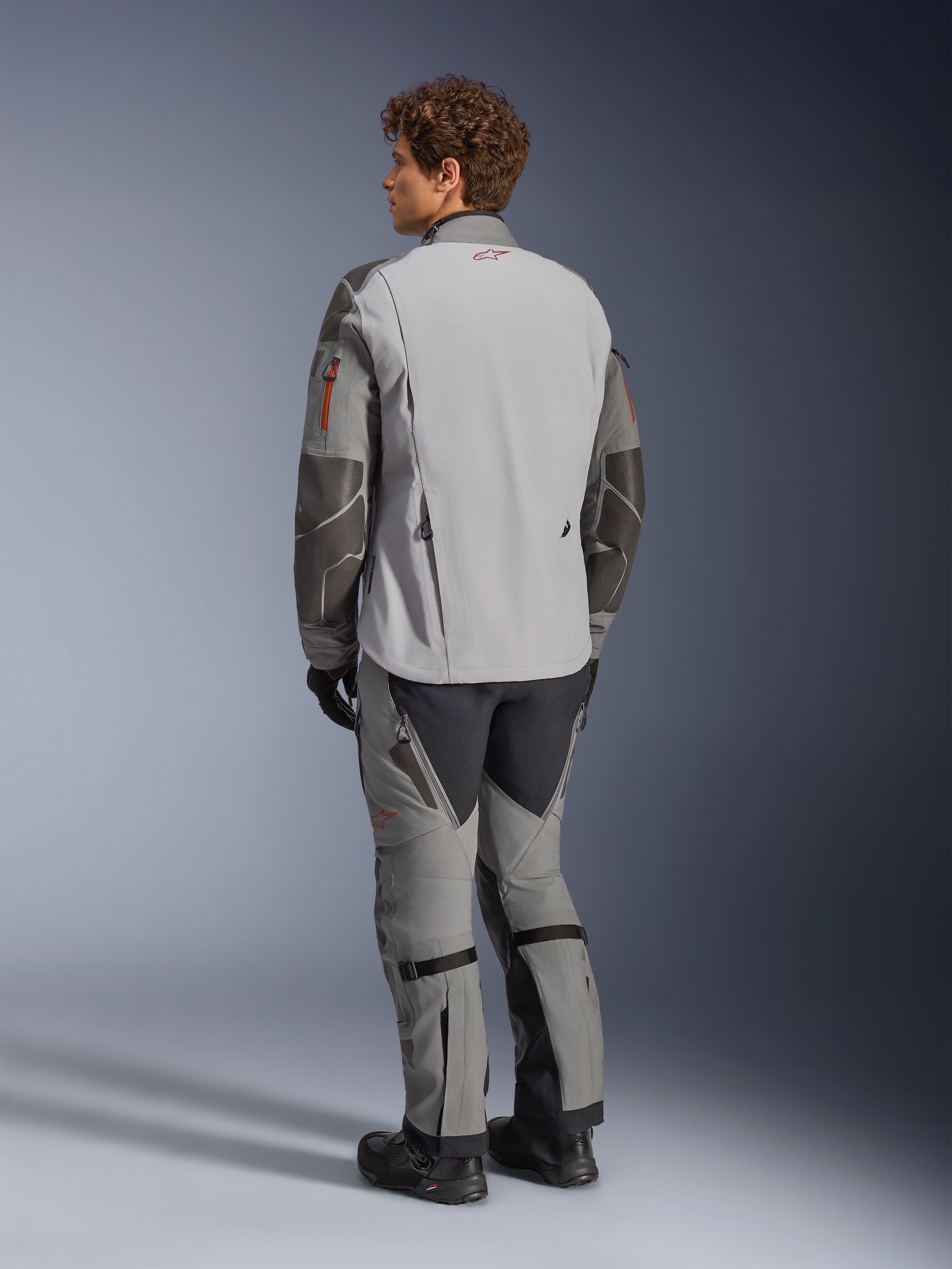 Alpinestars Nazca 3L GORE-TEX PRO Jacket, Adventure Motorcycle Jacket, Dark Gray and Mid Gray, rear view worn with matching adventure pants and black boots, featuring dark gray Superfabric reinforcements on the sleeves, orange zipper accents, and a small red logo on the back collar.
