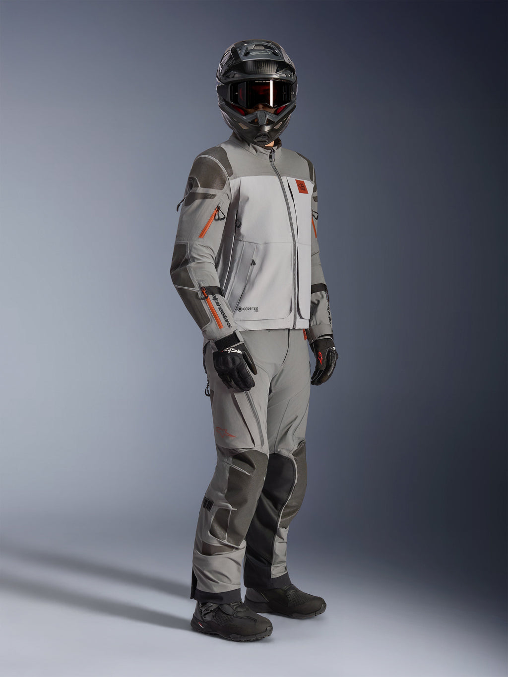 Alpinestars Nazca 3L GORE-TEX PRO Jacket, Adventure Motorcycle Jacket, Dark Gray and Mid Gray, worn as a complete outfit with matching adventure pants, featuring orange accents and Superfabric reinforcements, includes black adventure helmet and gloves, technical apparel for touring and off-road riding