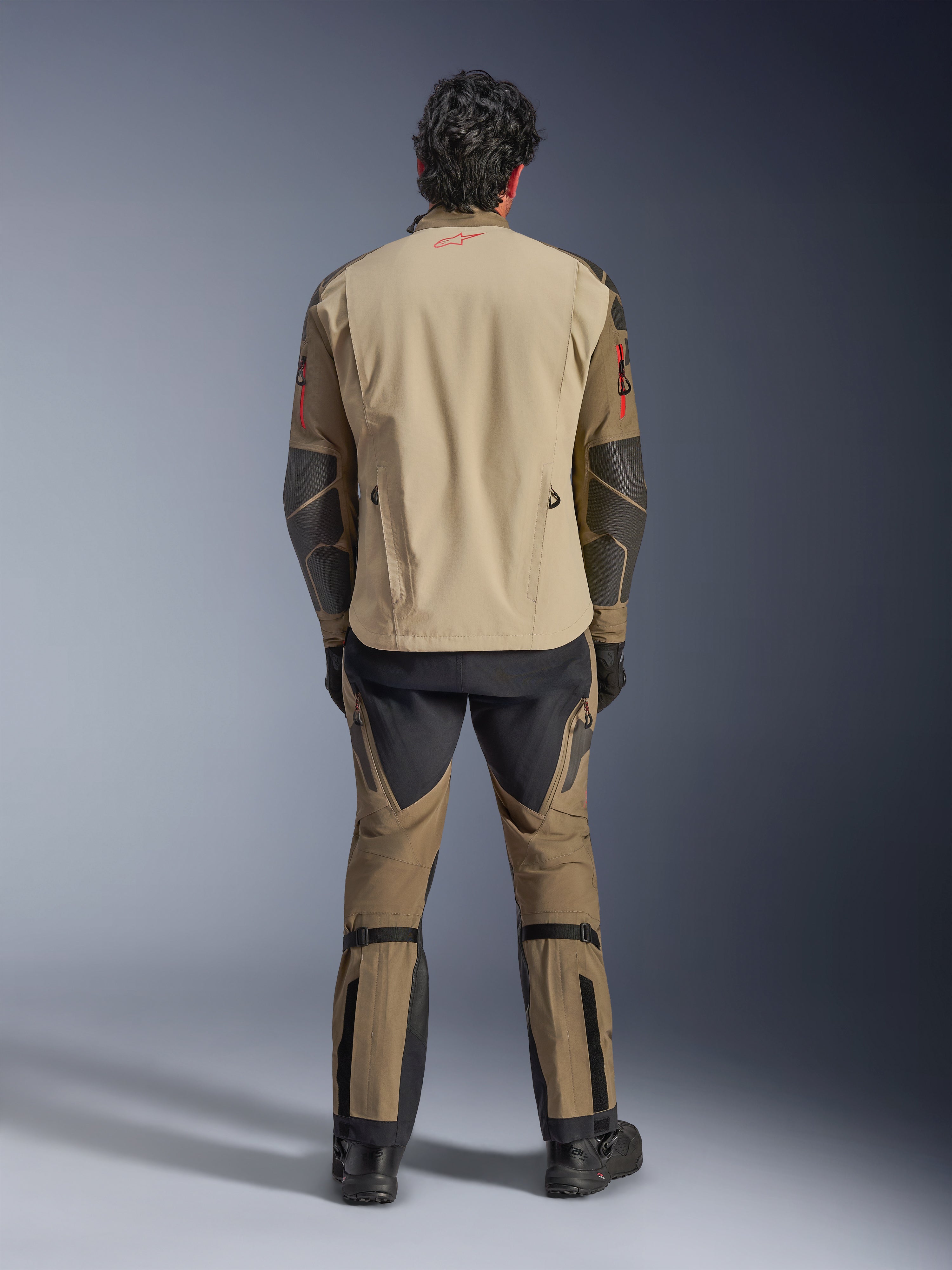Casaco Alpinestars Nazca 3L GORE-TEX PRO em Walnut Rock Brown, vista traseira do piloto, mostrando os reforços Superfabric nos cotovelos e fechos de ventilação traseiros, usado com calças de aventura a condizer, botas pretas e luvas, vestuário técnico de condução robusto para aventura extrema.