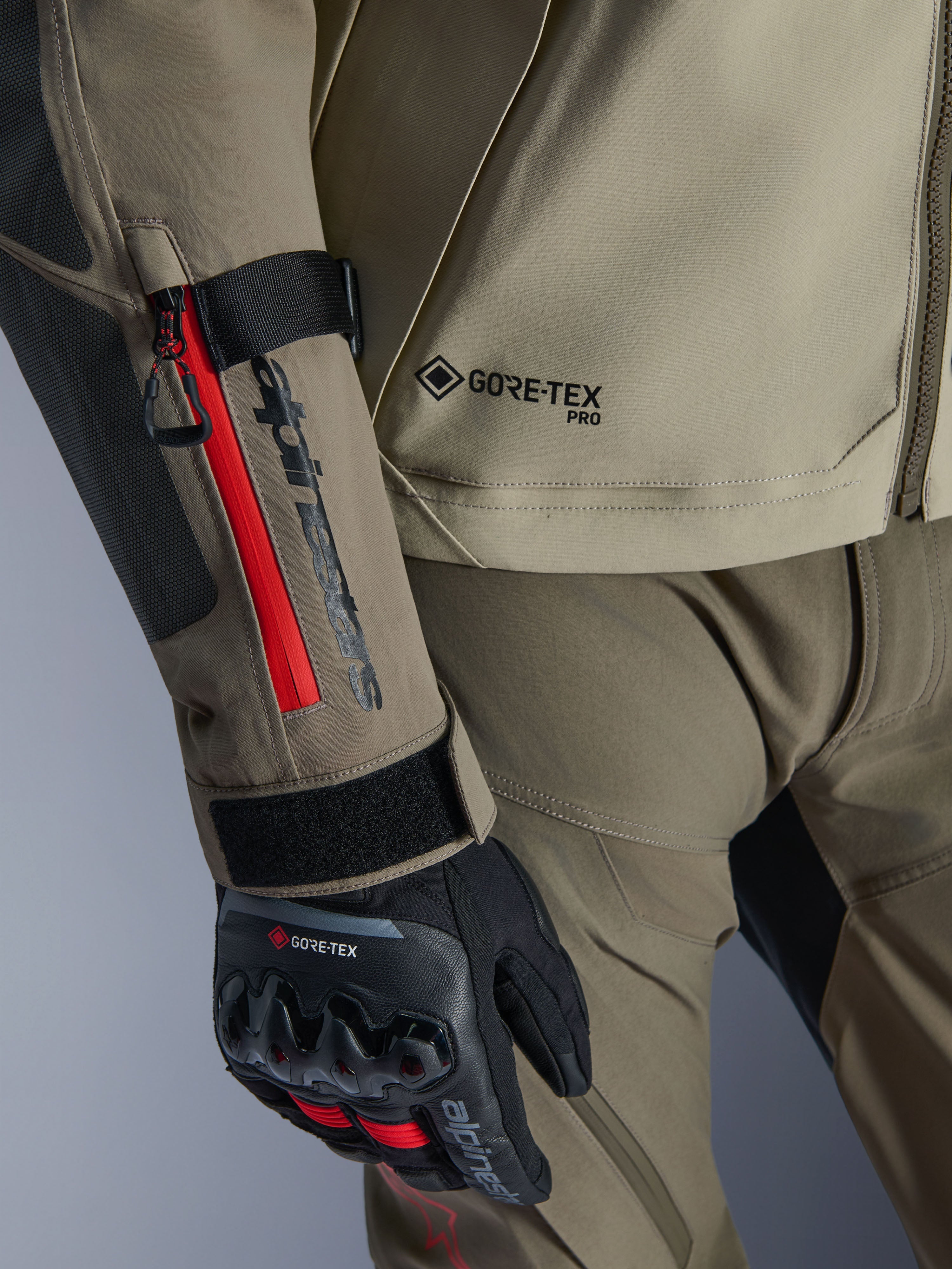 Pormenor do Casaco Alpinestars Nazca 3L GORE-TEX PRO em Walnut Rock Brown, com detalhes de fecho vermelhos na manga, logótipo GORE-TEX PRO e luvas de aventura pretas GORE-TEX