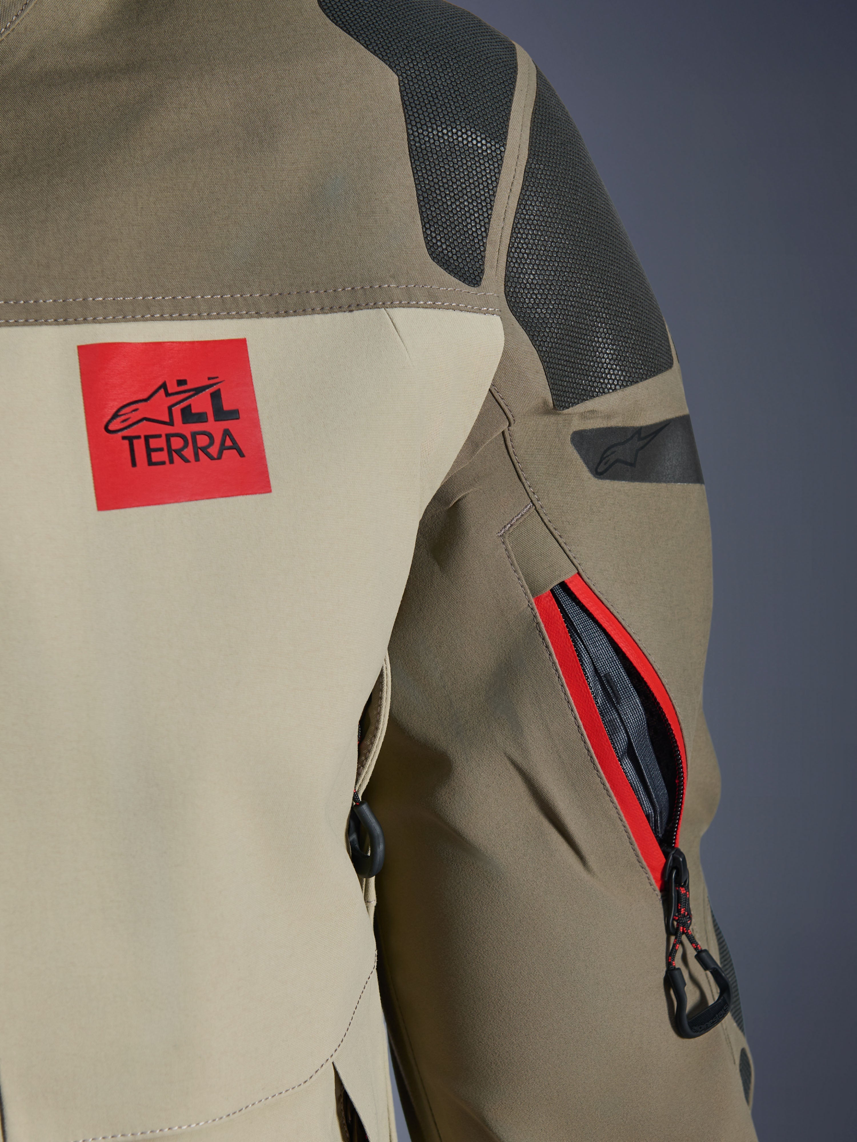 Casaco Alpinestars Nazca 3L GORE-TEX PRO, Walnut Rock Brown, pormenor da parte posterior do ombro e da manga, patch vermelho com o logótipo 'ALL TERRA', painéis de ombro em Superfabric preto texturizado, abertura de ventilação no braço com fecho de correr e contorno vermelho, vestuário de motociclismo de aventura premium.