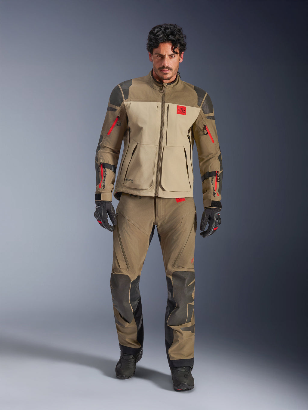 Casaco Alpinestars Nazca 3L GORE-TEX PRO, Walnut Rock Brown, casaco de motociclismo de aventura usado com calças e luvas a condizer, com detalhes de fechos de correr vermelhos, logótipo 'All Terra' no peito e construção têxtil robusta.