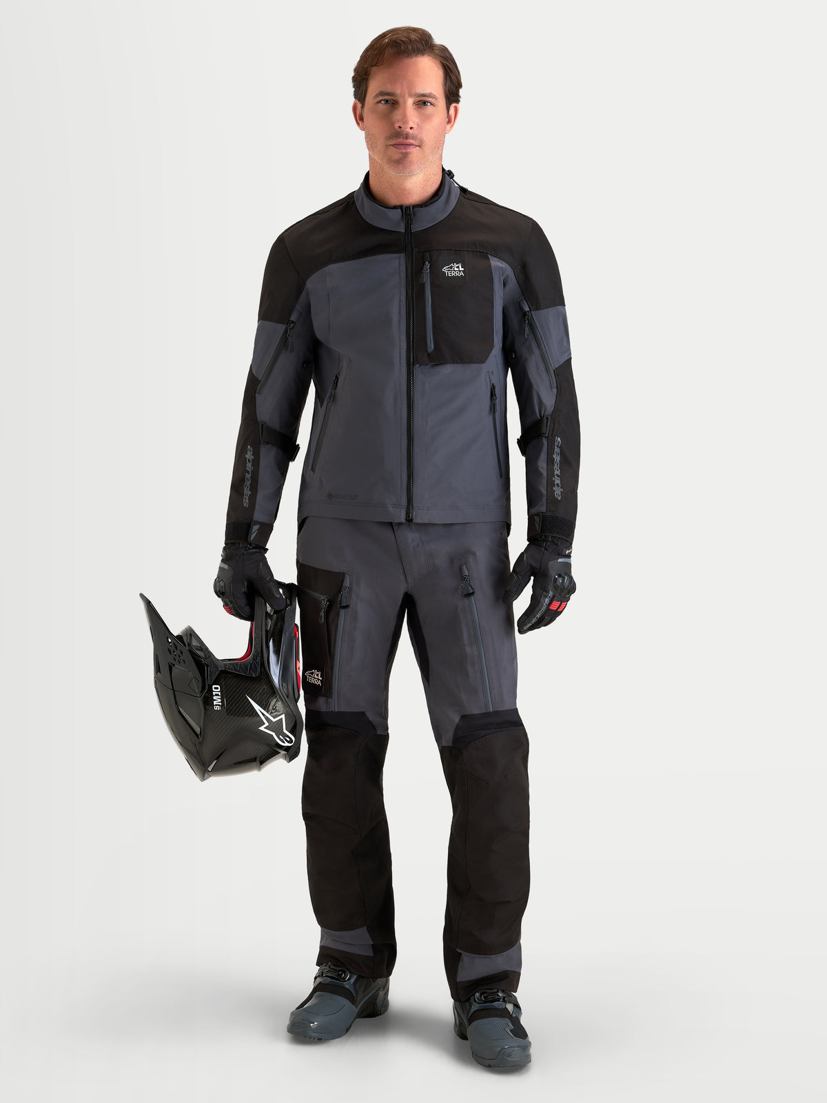 Alpinestars Arenis Gore-Tex casaco, casaco de moto adventure para homem em preto e cinzento escuro, vestido por um condutor a segurar um capacete, destacando a construção em tecido impermeável GORE-TEX