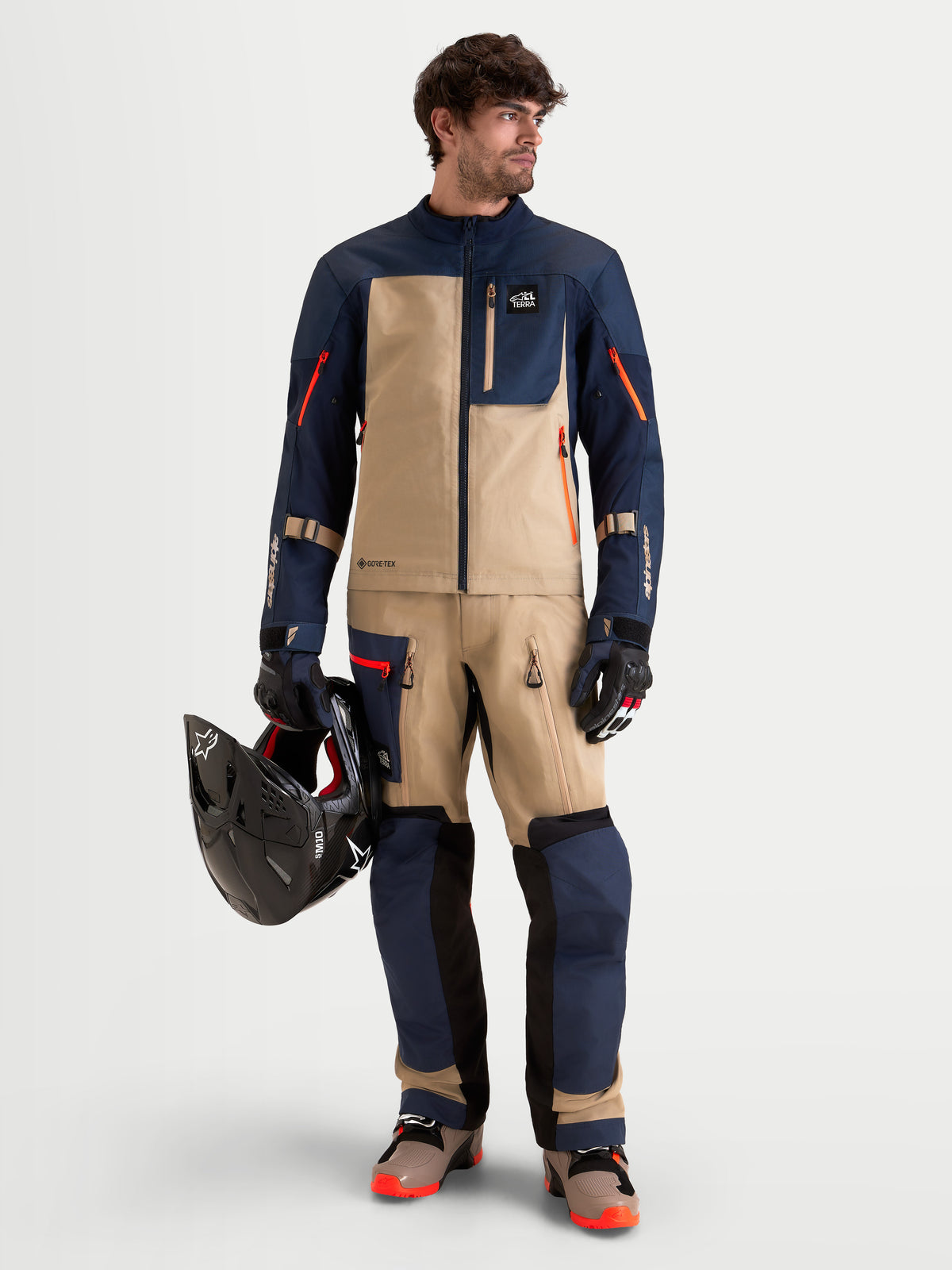 Alpinestars Arenis Gore-Tex Gear Set, conjunto de equipamento de aventura para homem em azul-marinho, bege e preto, vista frontal num modelo a vestir o blusão e as calças a condizer e a segurar um capacete, equipamento impermeável certificado AA