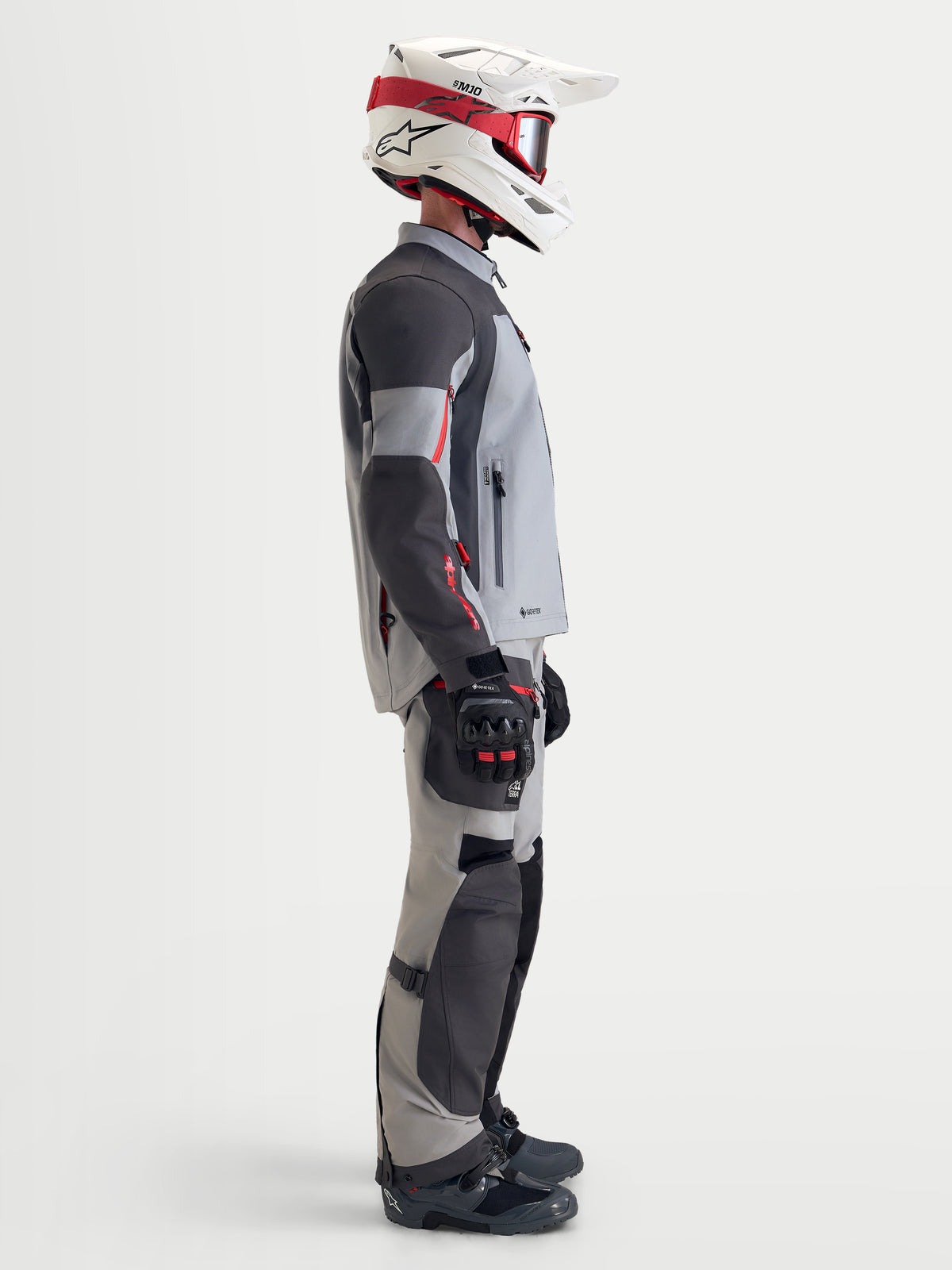 Alpinestars Arenis Gore-Tex, casaco de moto adventure para homem em cinzento-médio, cinzento-escuro e preto, vista de perfil lateral de um motociclista a usar o blusão têxtil impermeável com equipamento a condizer e um capacete branco e vermelho