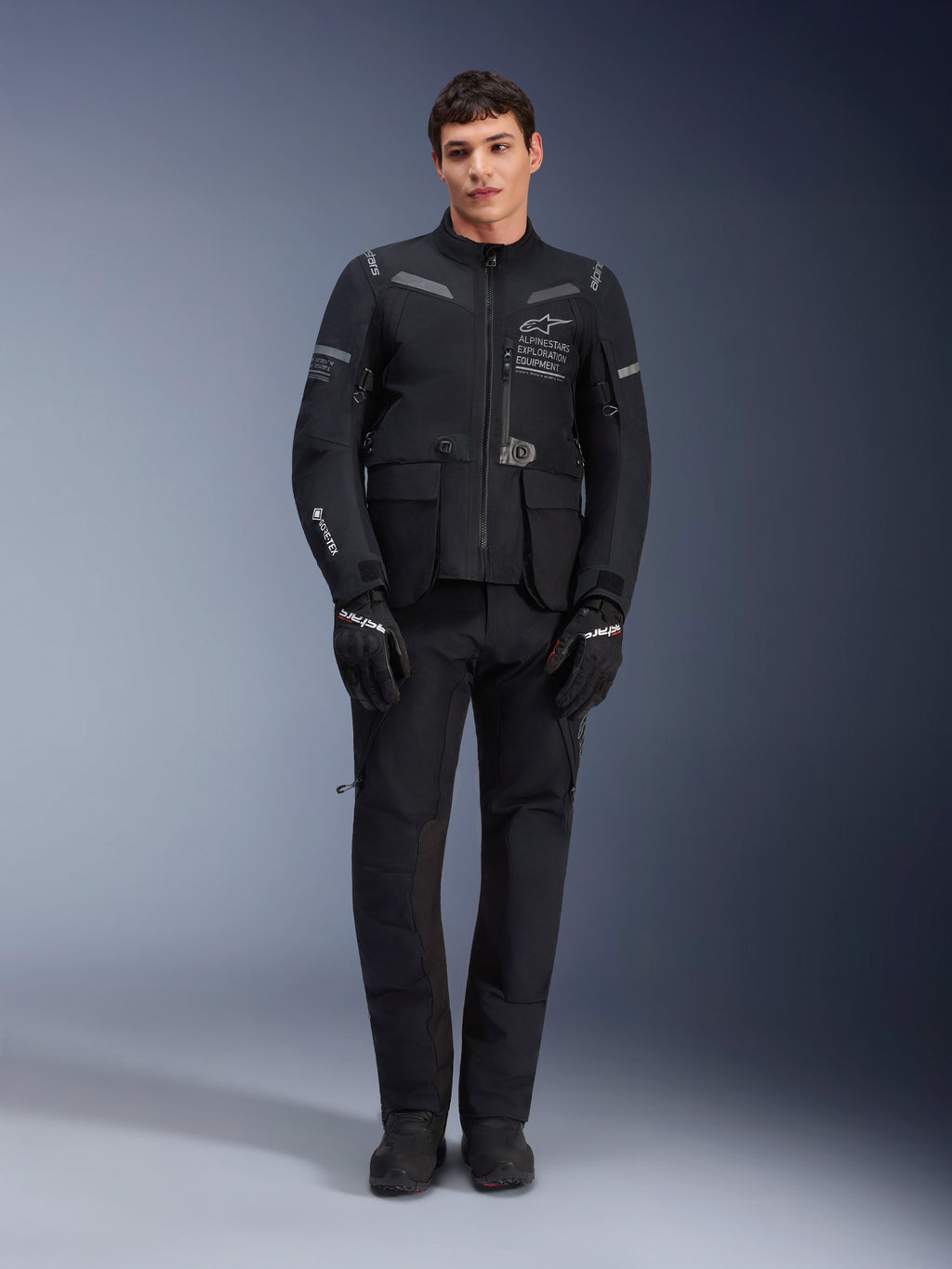 Casaco Alpinestars ST-7 2L GORE-TEX, casaco de motociclismo adventure touring, preto e cinzento escuro, usado com calças e luvas de motociclismo pretas a condizer, com vários bolsos de carga, detalhes refletores e logótipo Alpinestars Exploration Equipment no peito, design funcional para condução em estrada e fora de estrada