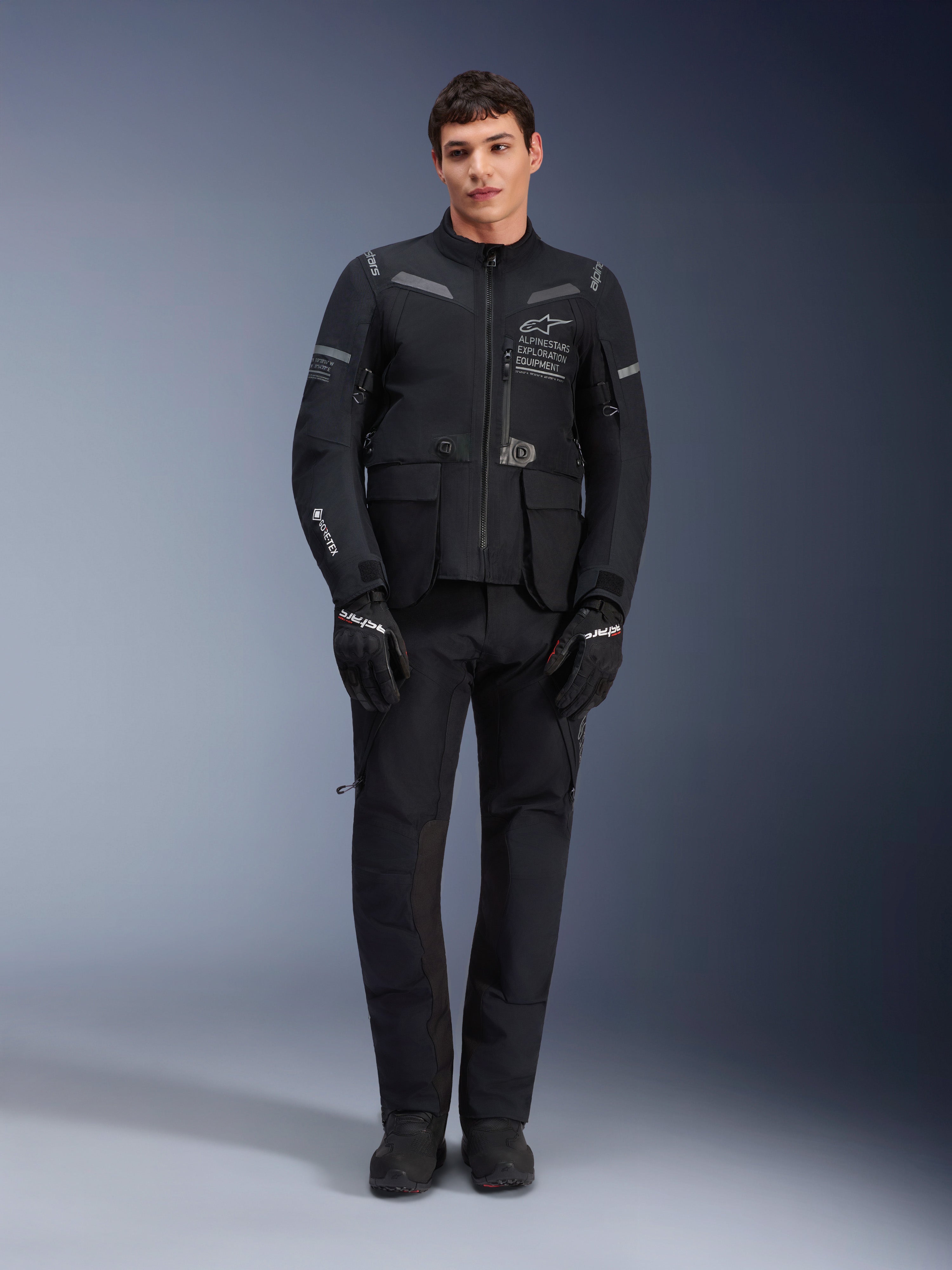 Casaco Alpinestars ST-7 2L GORE-TEX, casaco de motociclismo adventure touring, preto e cinzento escuro, usado com calças e luvas de motociclismo pretas a condizer, com vários bolsos de carga, detalhes refletores e logótipo Alpinestars Exploration Equipment no peito, design funcional para condução em estrada e fora de estrada