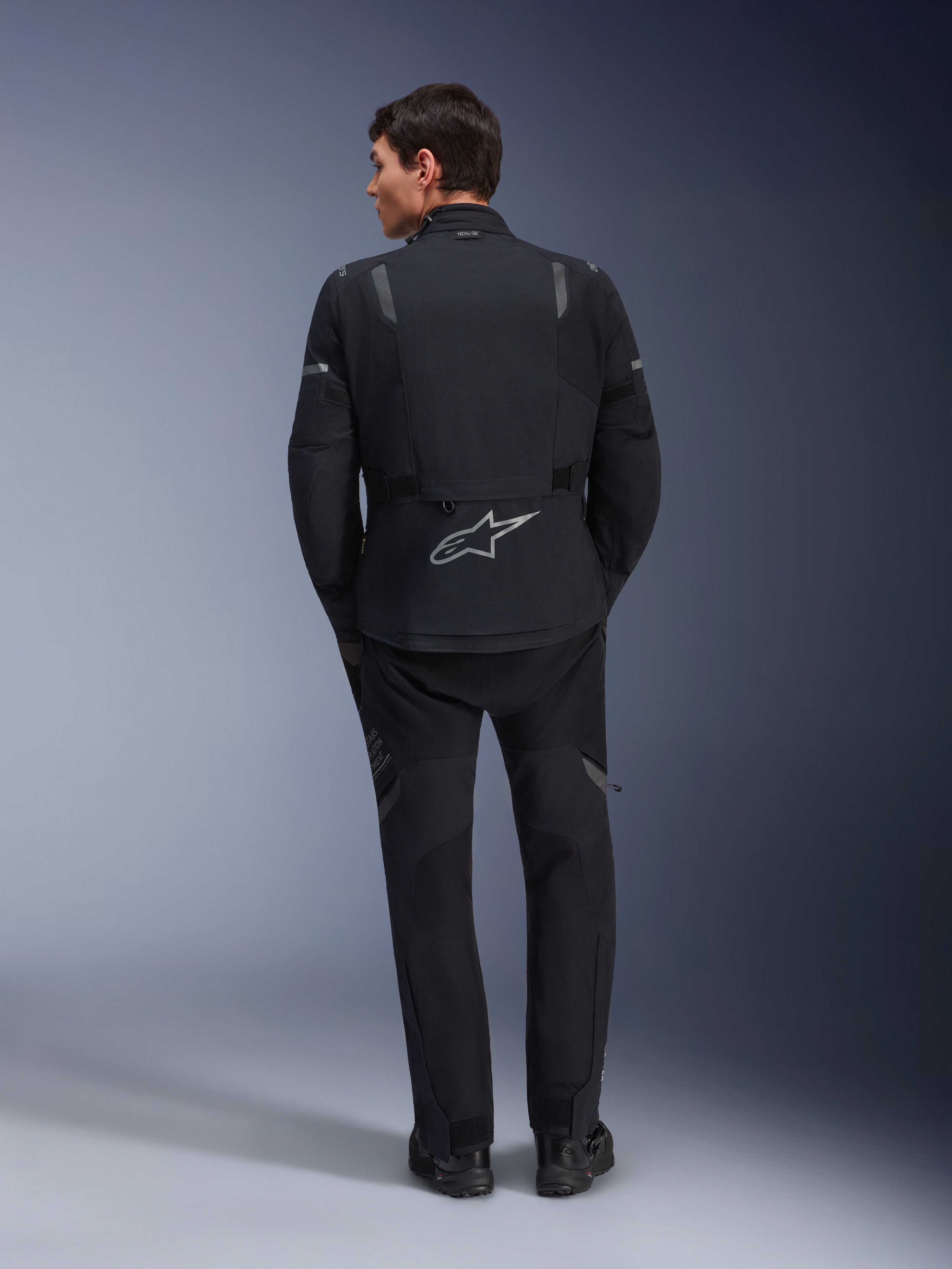 Casaco Alpinestars ST-7 2L GORE-TEX e calças ST-7 2L GORE-TEX a condizer, equipamento de motociclismo de aventura em preto e cinzento escuro, vista traseira com logótipo Alpinestars refletor no casaco e construção têxtil técnica.