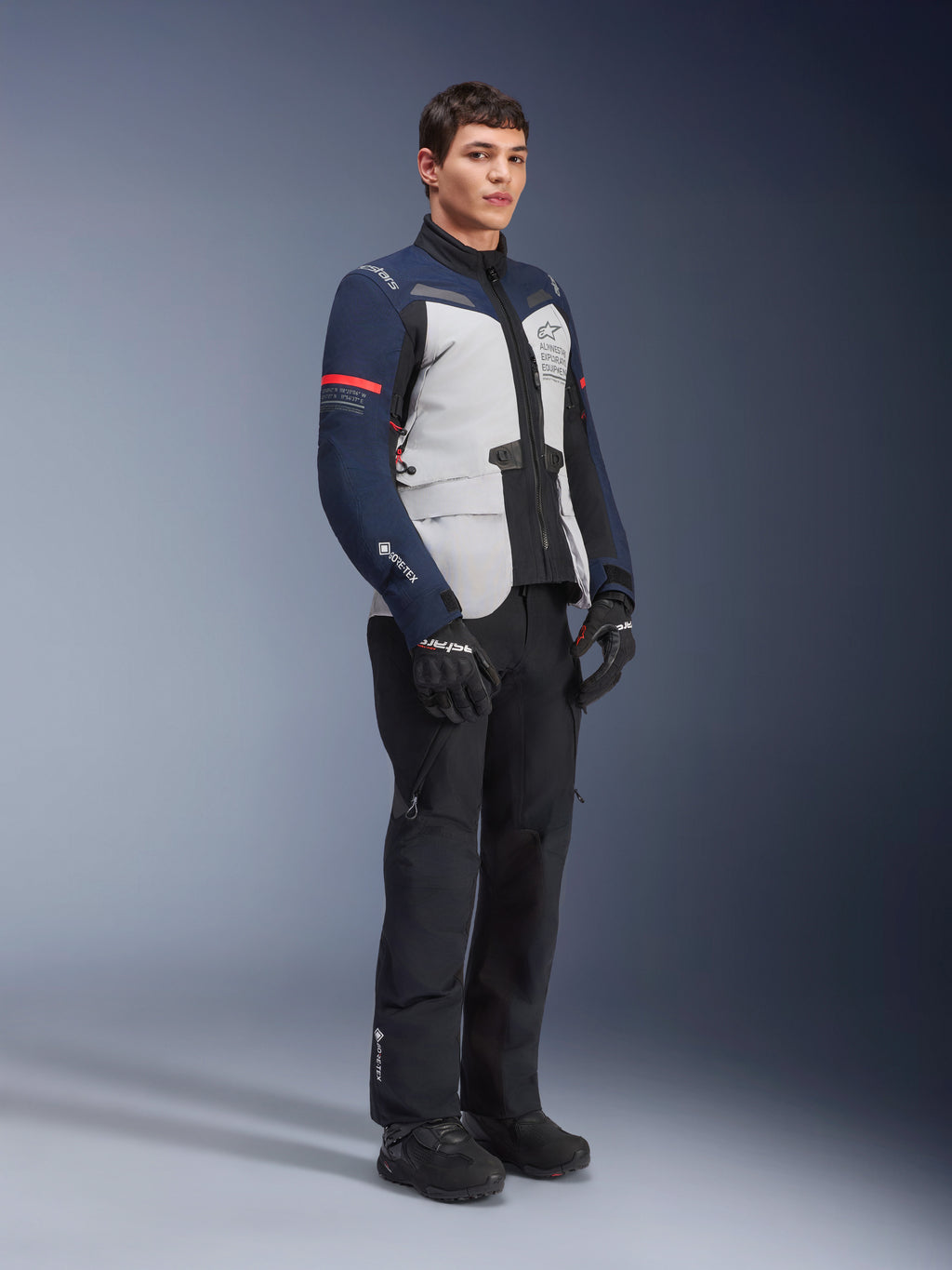 Alpinestars ST-7 2L GORE-TEX Jacket in Ice Gray, Dark Blue, and Black colorway with red accents, worn by a male model with black motorcycle pants and gloves, professional adventure touring motorcycle gear