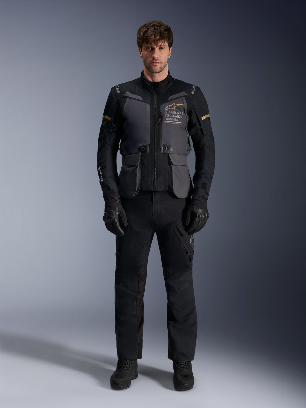 Male model wearing the Alpinestars ST-7 2L GORE-TEX Jacket in Deep Gray Black Dark Khaki, featuring 'ALPINESTARS EXPLORATION EQUIPMENT' branding on the chest, paired with matching black riding pants, gloves, and boots, studio front view.