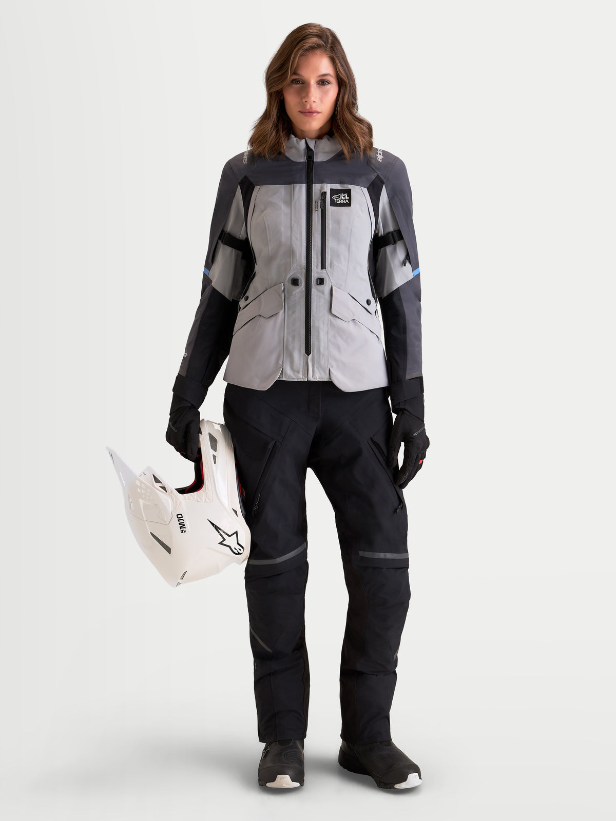 Alpinestars Stella ST-7 V2 2L Gore-Tex Jacket, women's all-terrain adventure touring motorcycle jacket in mid gray, deep gray, and black, front view worn by a model holding a white off-road helmet, featuring waterproof textile construction and CE certification.