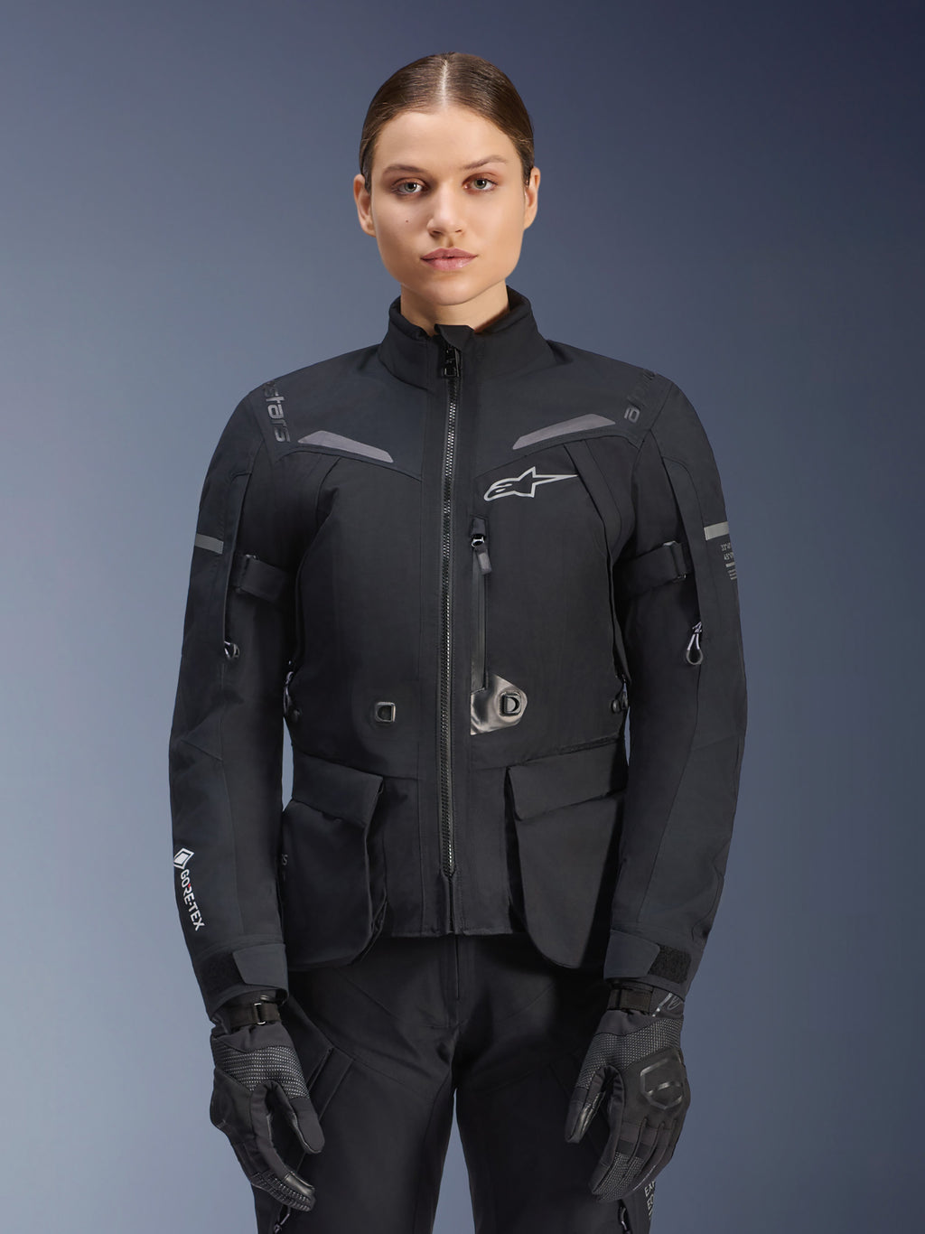 Mulher a usar o casaco Alpinestars Stella ST-7 2L GORE-TEX, preto com detalhes em cinzento escuro, casaco de touring para motociclismo com bolsos de carga grandes e gola alta, combinado com luvas e calças pretas, vestuário de aventura impermeável