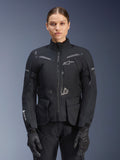 Mulher a usar o casaco Alpinestars Stella ST-7 2L GORE-TEX, preto com detalhes em cinzento escuro, casaco de touring para motociclismo com bolsos de carga grandes e gola alta, combinado com luvas e calças pretas, vestuário de aventura impermeável