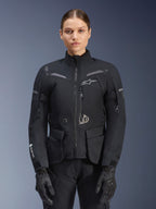 Mulher a usar o casaco Alpinestars Stella ST-7 2L GORE-TEX, preto com detalhes em cinzento escuro, casaco de touring para motociclismo com bolsos de carga grandes e gola alta, combinado com luvas e calças pretas, vestuário de aventura impermeável