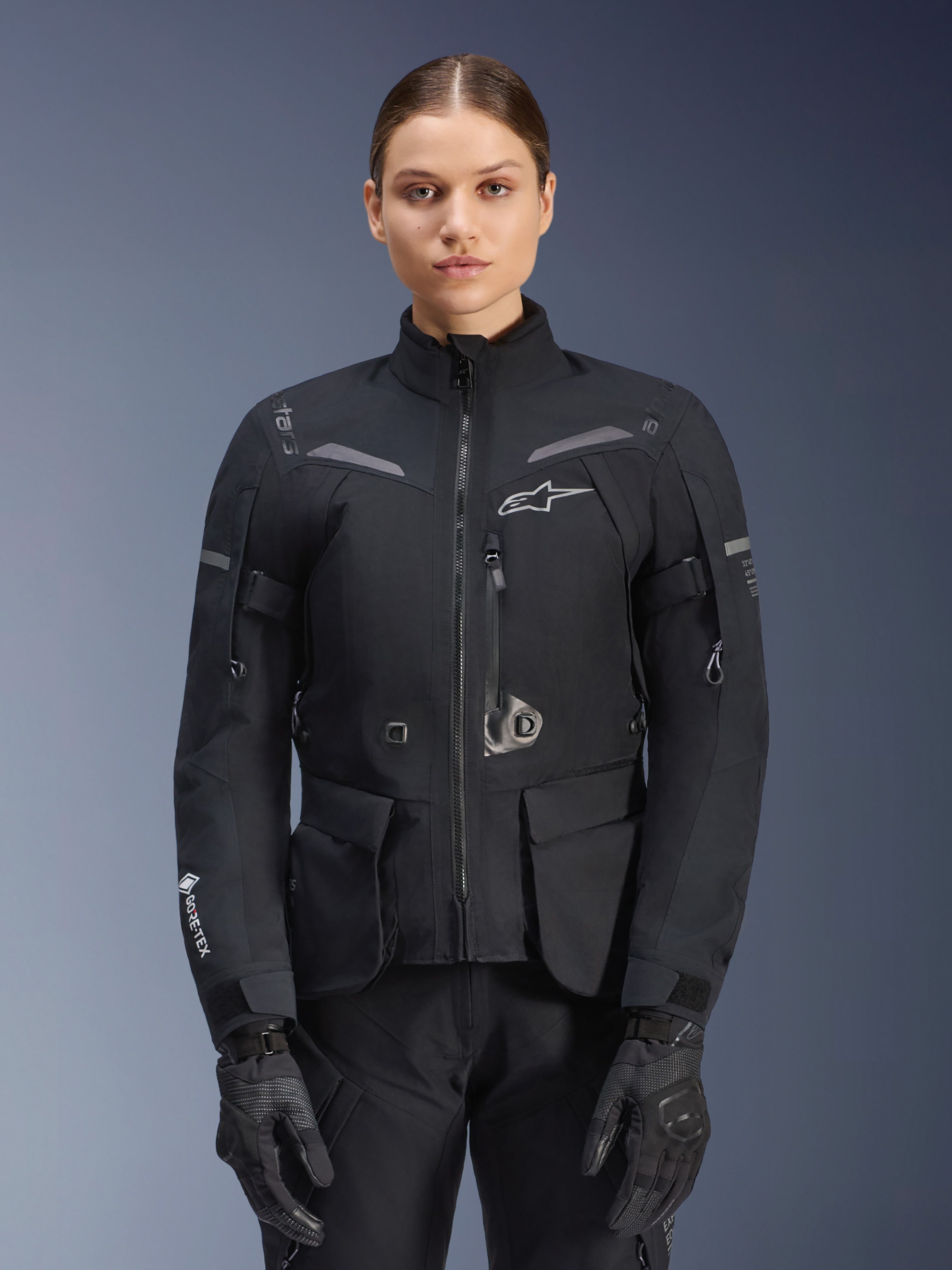 Mulher a usar o casaco Alpinestars Stella ST-7 2L GORE-TEX, preto com detalhes em cinzento escuro, casaco de touring para motociclismo com bolsos de carga grandes e gola alta, combinado com luvas e calças pretas, vestuário de aventura impermeável