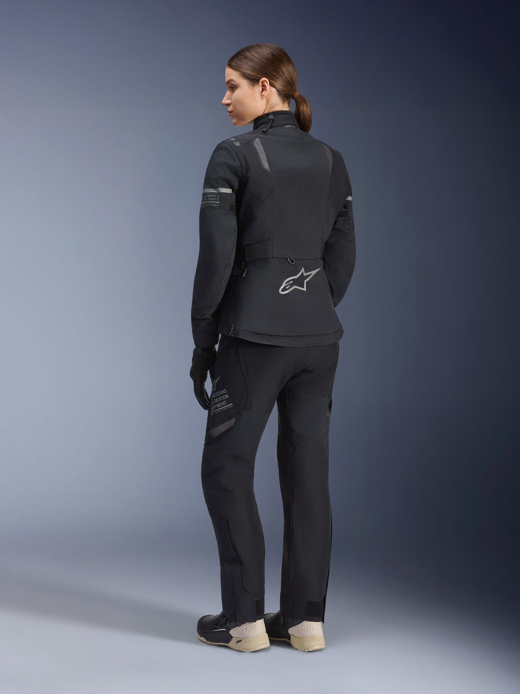 Jaqueta Stella ST-7 2L GORE-TEX para Mulher, Preto e Cinzento Escuro, jaqueta de turismo de aventura vista de costas, usada com calças pretas a condizer e botas creme, com o logótipo Alpinestars na zona lombar, concebida para touring de estrada em todas as estações