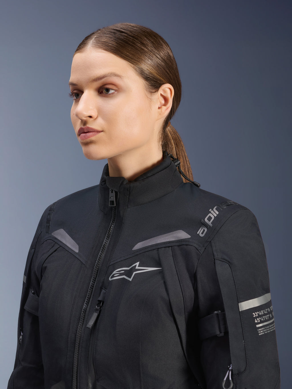 Jaqueta Stella ST-7 2L GORE-TEX Feminina da Alpinestars, preta, com logótipo no peito e coordenadas técnicas no braço, design têxtil impermeável de gola alta para touring de motociclismo, retrato de estúdio de uma motociclista.