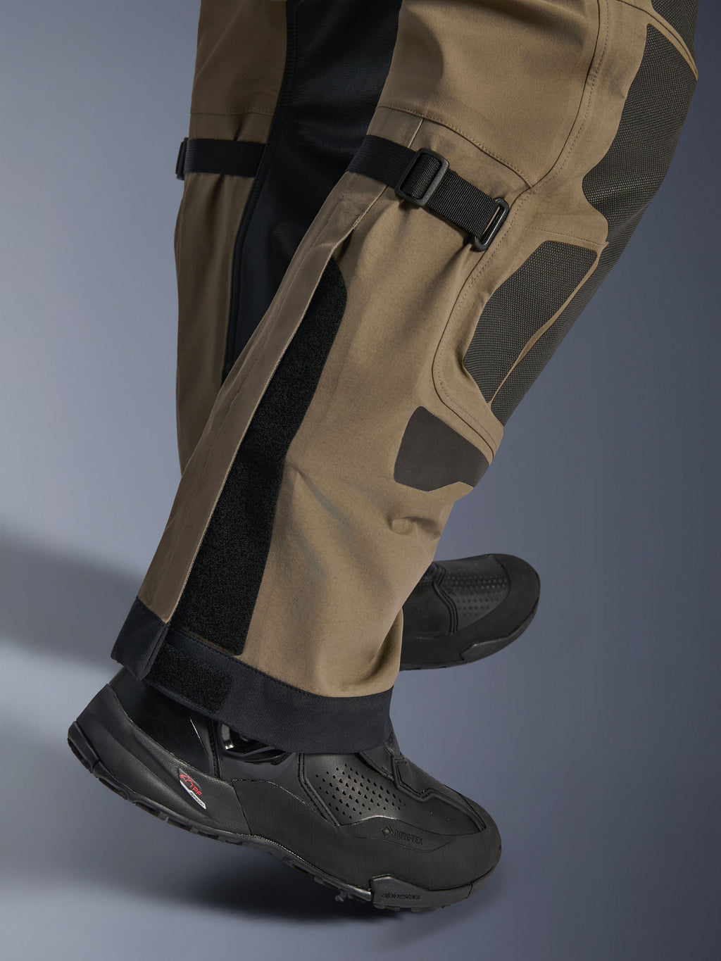 Pormenor das calças Alpinestars Nazca 3L GORE-TEX PRO em Walnut Black, mostrando detalhes técnicos da parte inferior da perna, incluindo ajustadores de volume na panturrilha e reforços Superfabric, usadas com botas de motociclismo de aventura pretas.