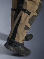 Pormenor das calças Alpinestars Nazca 3L GORE-TEX PRO em Walnut Black, mostrando detalhes técnicos da parte inferior da perna, incluindo ajustadores de volume na panturrilha e reforços Superfabric, usadas com botas de motociclismo de aventura pretas.