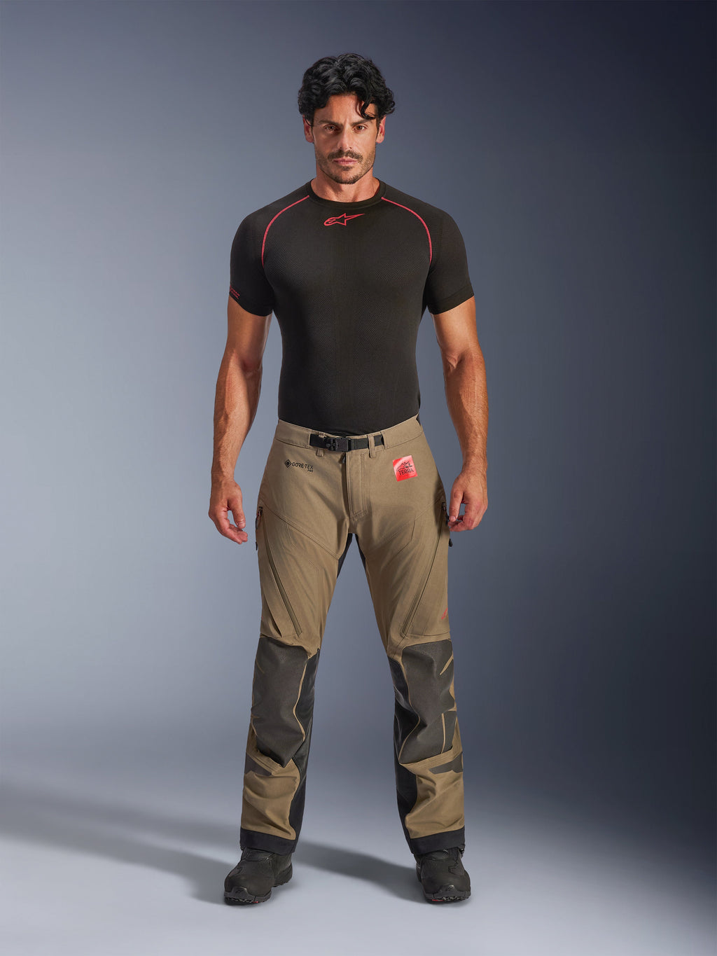 Modelo masculino a vestir Calças Alpinestars Nazca 3L GORE-TEX PRO em castanho nogueira e preto, equipamento técnico de aventura com reforços de joelho em Superfabric e fivela magnética na cintura, combinado com uma camisola de base preta Alpinestars, vista frontal de estúdio.