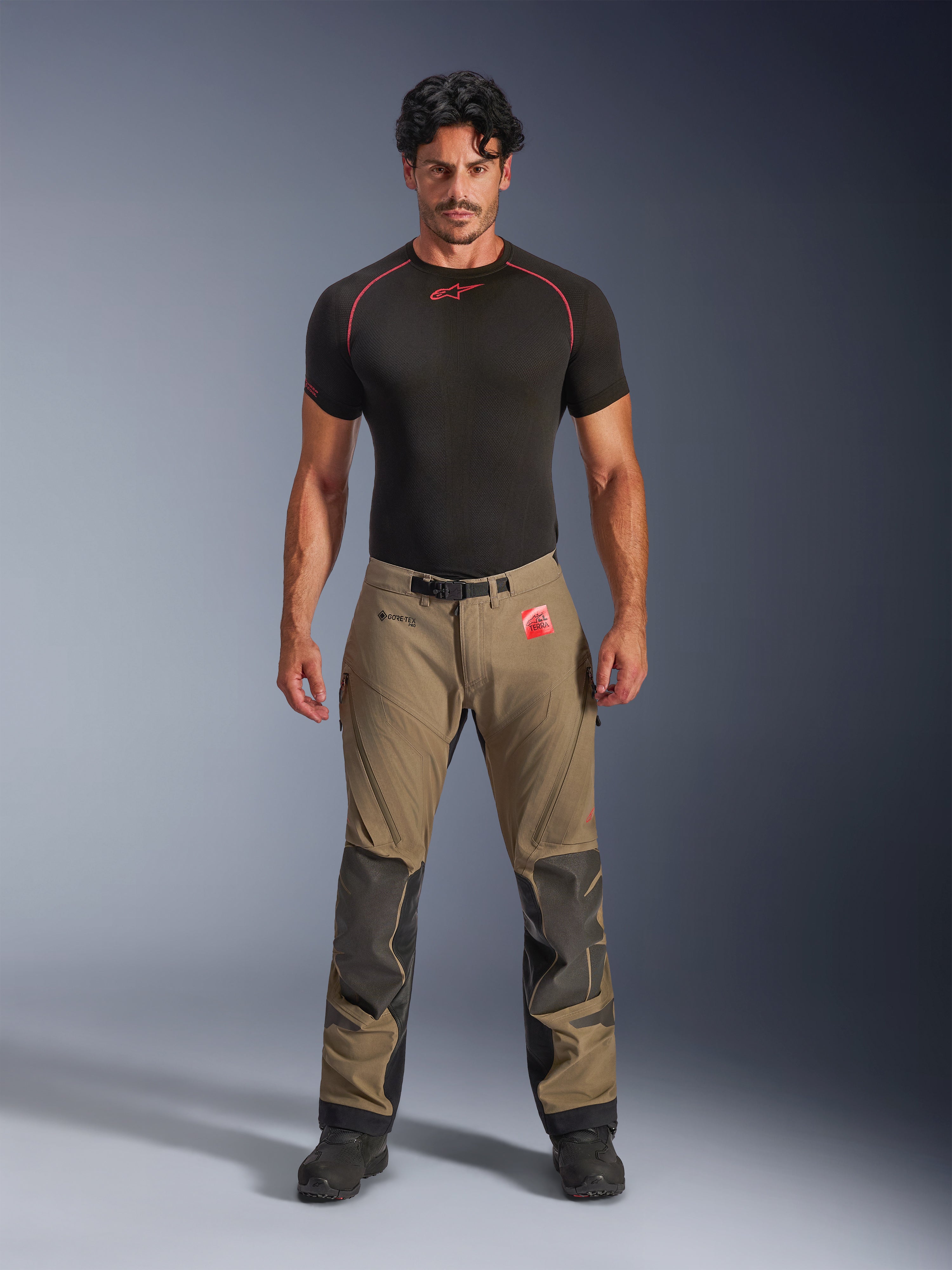 Modelo masculino a vestir Calças Alpinestars Nazca 3L GORE-TEX PRO em castanho nogueira e preto, equipamento técnico de aventura com reforços de joelho em Superfabric e fivela magnética na cintura, combinado com uma camisola de base preta Alpinestars, vista frontal de estúdio.