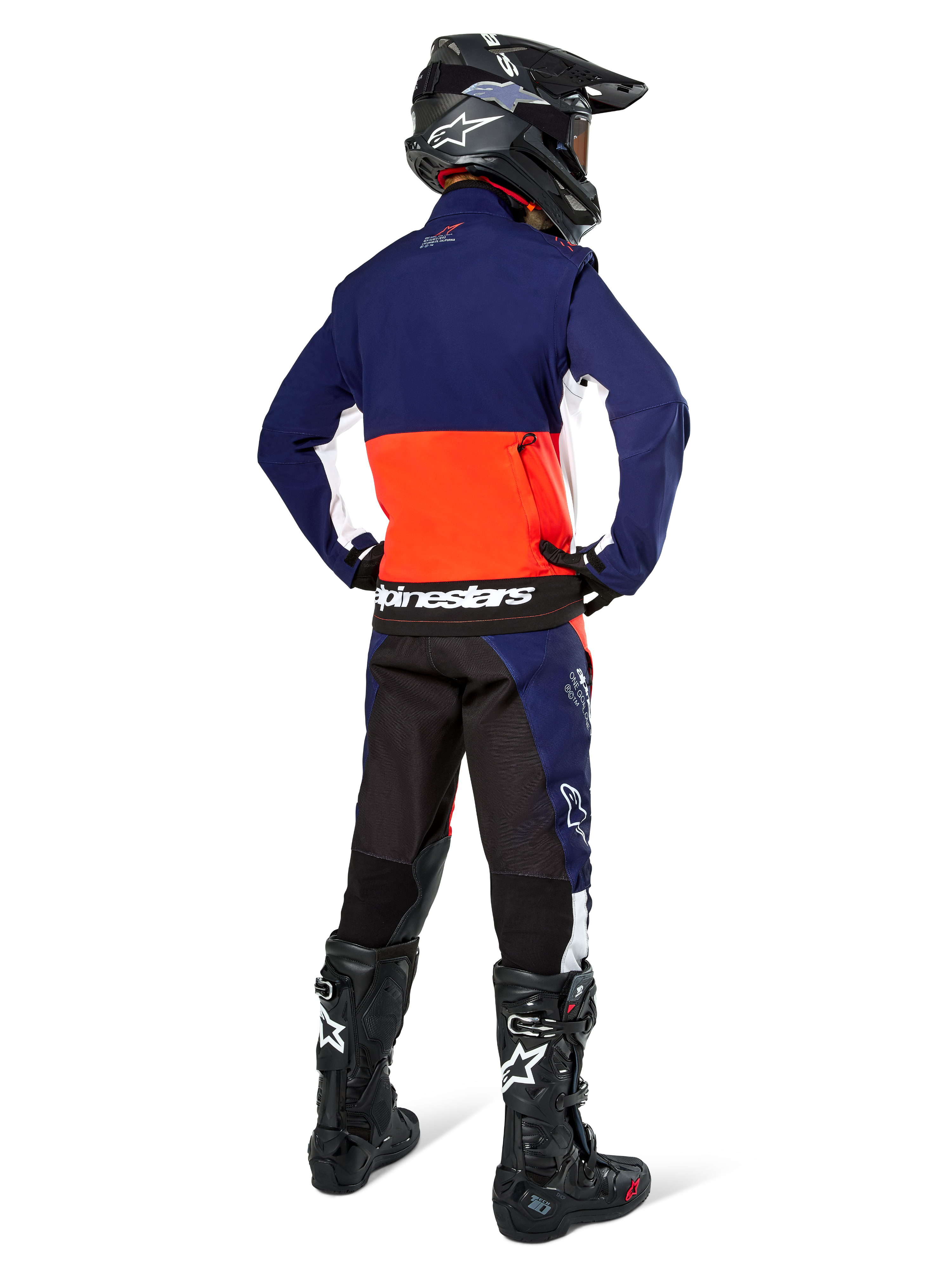 Motocross rider from the back wearing Alpinestars Lite-Dura Softshell Jacket in Navy, Hot Orange, and White, showing the large rear storage pocket. Styled with matching enduro pants, a black motocross helmet, and black off-road boots.