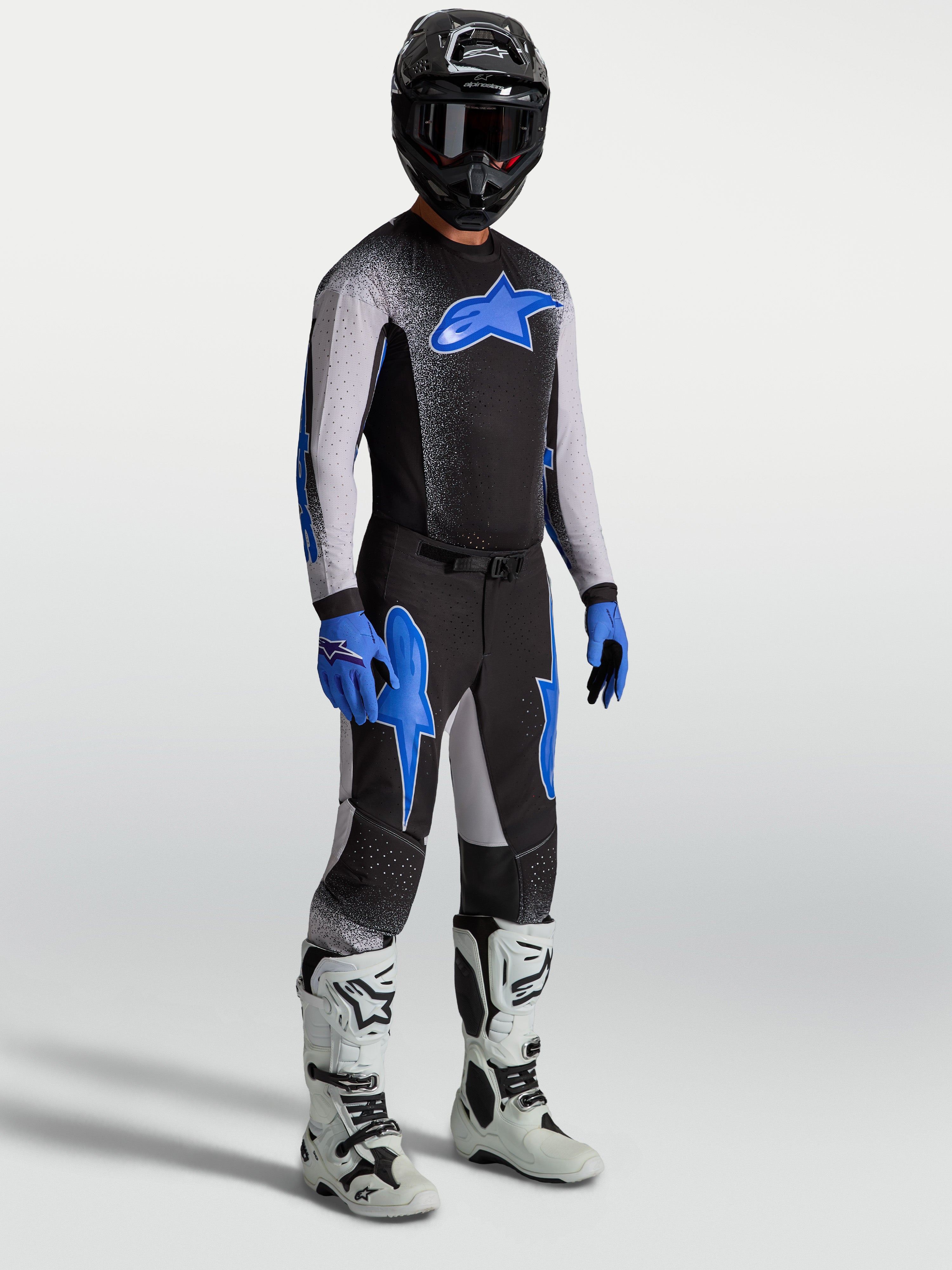 Alpinestars Supertech Hiten AFD, calças de motocross para homem, pretas e azuis, em modelo com camisola e botas brancas.