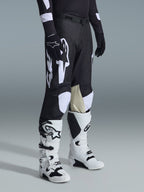 Alpinestars 2026 Racer Air Riway Pants, Black and White motocross pants with large white logo graphics, beige leather inner knee panels, worn with white and black off-road boots and black gloves in a studio setting.