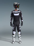 Piloto de motocross com calças Alpinestars 2026 Racer Air Riway em preto e branco, camisola preta e branca a condizer com o logótipo 'astars', capacete preto, óculos com lentes iridescentes e botas de corrida brancas, conjunto de equipamento profissional para todo-o-terreno