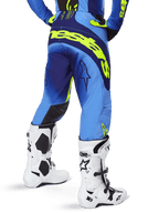 Alpinestars 2025 Techstar Melt Pants, calças de motocross, Amarelo Fluo Azul, vista traseira com logótipo amarelo grande na zona do assento e detalhes em azul escuro, usadas com botas brancas Alpinestars Tech 10 e camisola a condizer.
