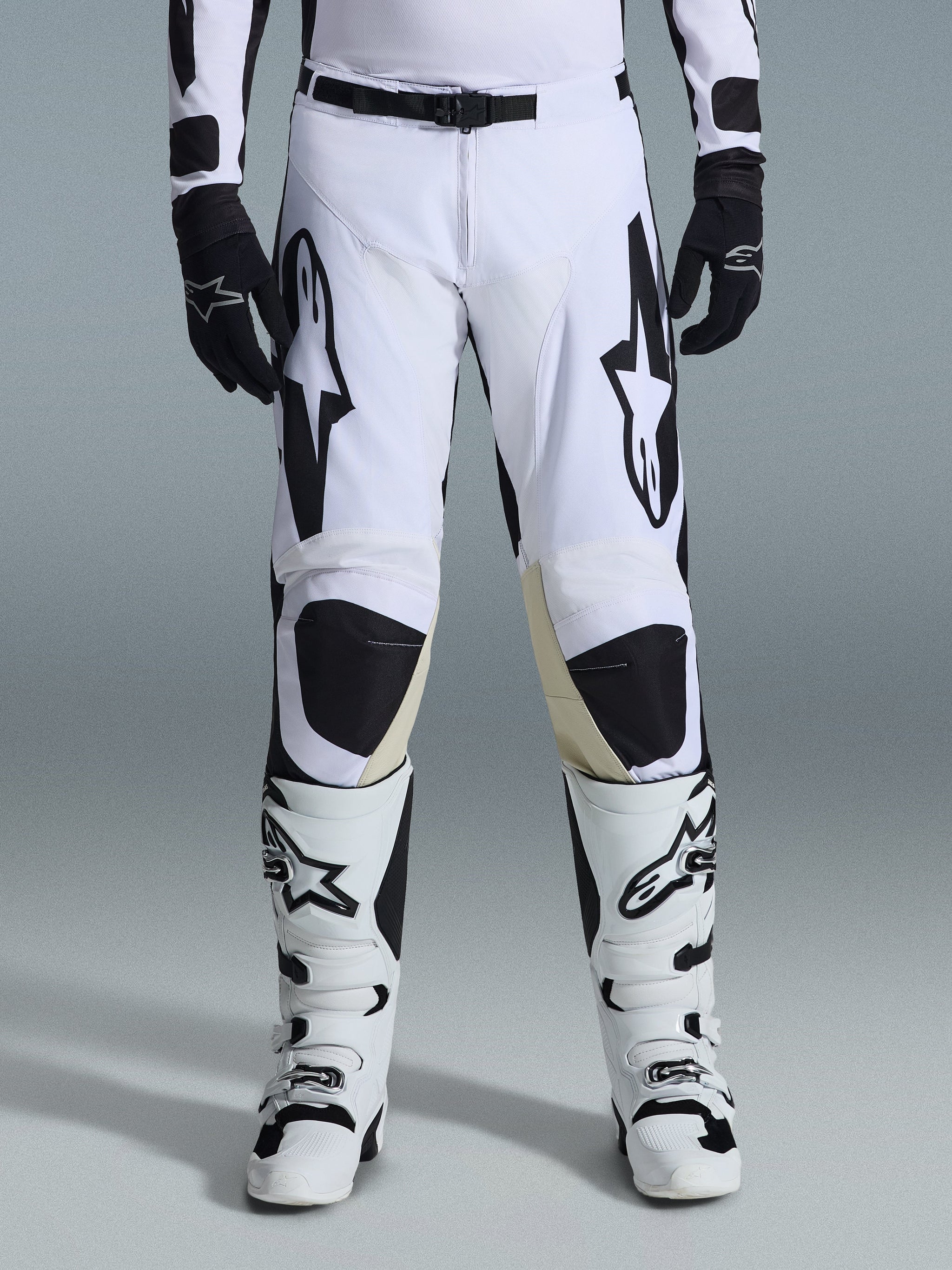 Calças Alpinestars Racer Riway 2026, calças de motocross, em branco e preto, usadas com botas de motocross brancas e pretas a condizer e luvas pretas, com logótipos Alpinestars pretos de destaque nas coxas e design técnico para performance off-road