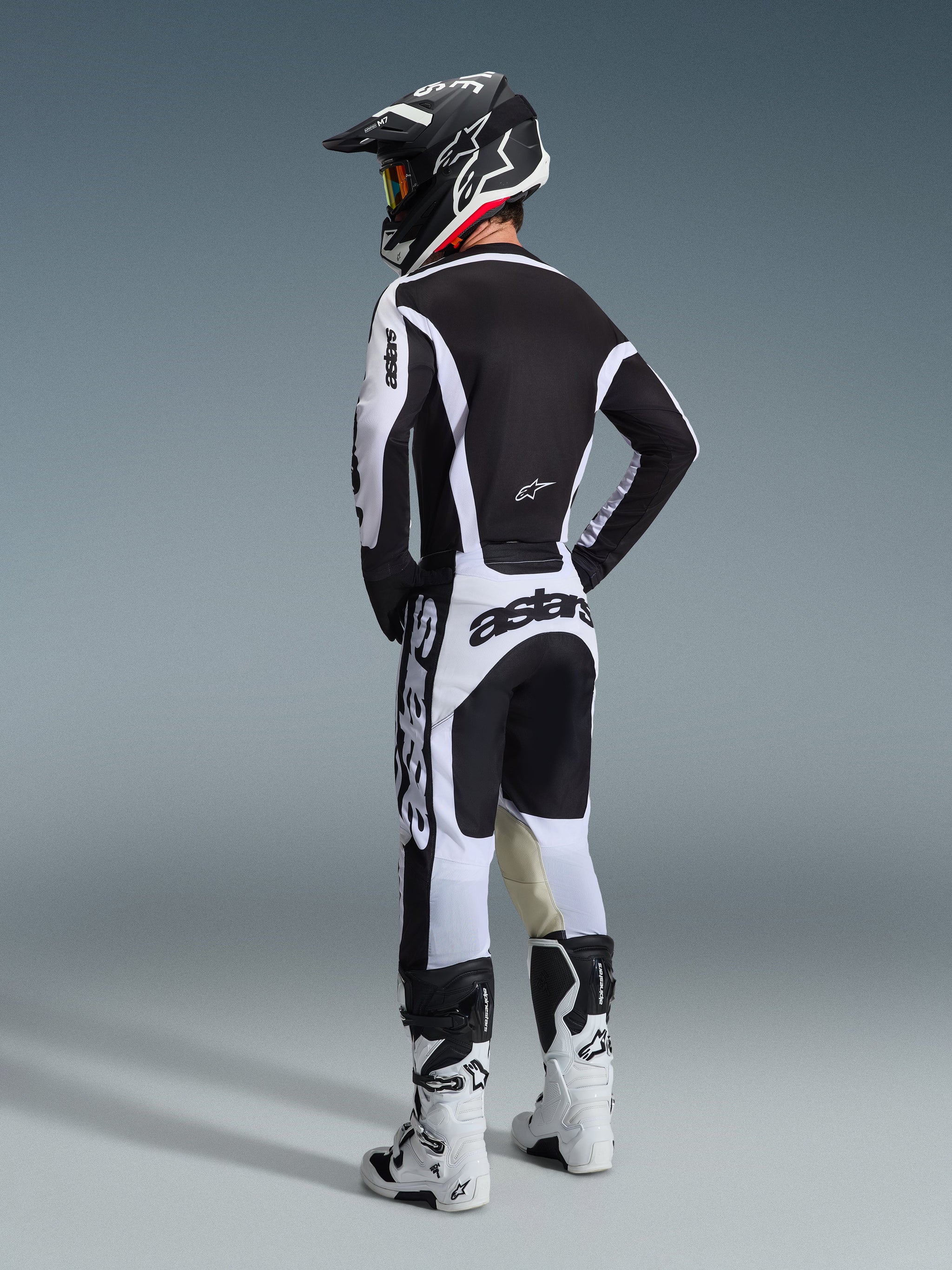 Piloto de motocross visto de trás a usar Calças Alpinestars 2026 Racer Riway em Branco e Preto, camisola preta e branca a condizer com logótipos destacados, capacete preto e branco e botas de motocross brancas, conjunto completo de equipamento de corrida para performance off-road.
