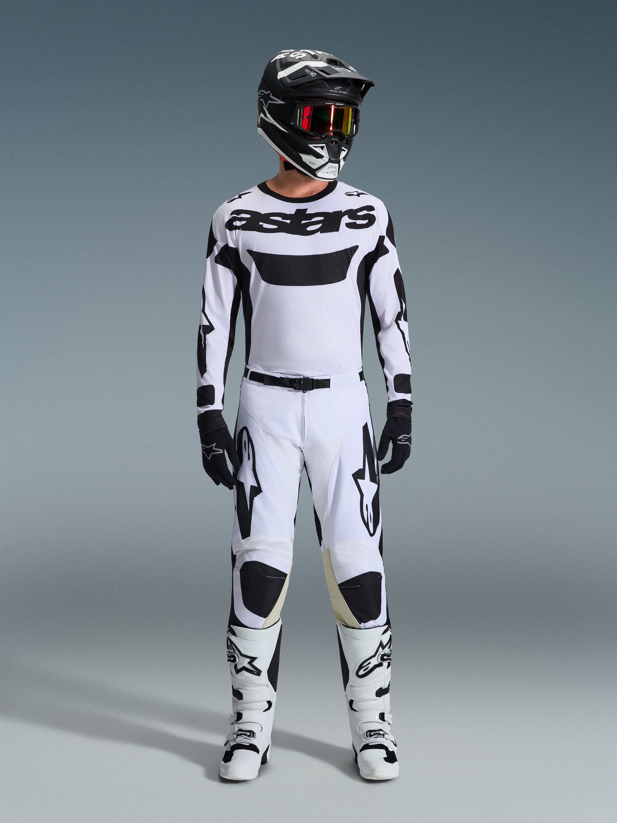 Piloto de motocross com calças Alpinestars 2026 Racer Riway em branco e preto, camisola branca a condizer, capacete preto com óculos vermelhos, luvas pretas e botas brancas, com logótipo Astars preto em destaque e um design focado no desempenho para uso off-road.