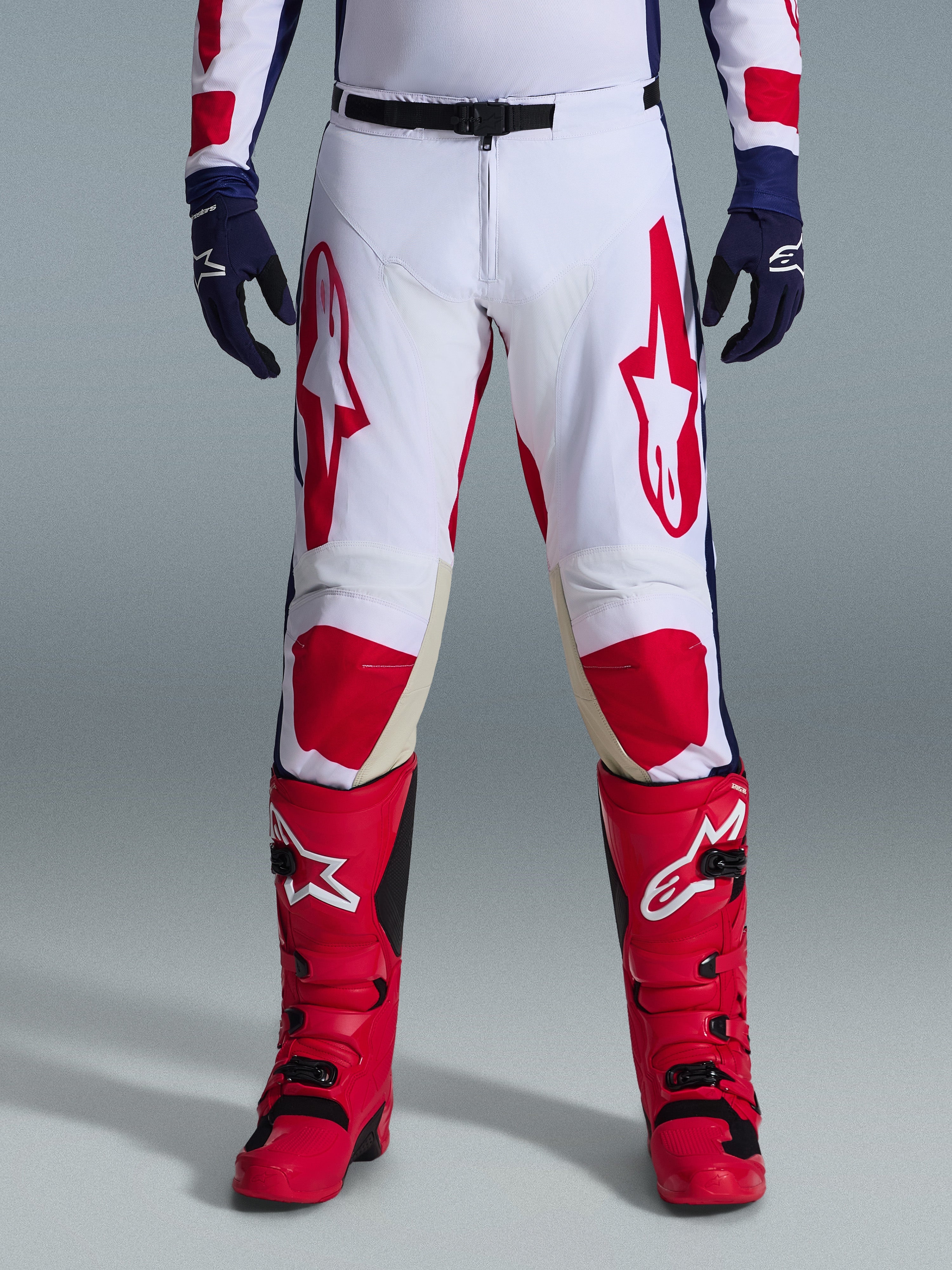 Alpinestars 2026 Racer Riway Pants, calças de motocross, brancas, azuis e vermelhas, usadas com botas vermelhas e luvas azul-escuro, com logótipos vermelhos grandes nas coxas e painéis vermelhos nos joelhos, design desportivo pré-curvado com fivela de ajuste na cintura