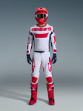 Piloto de motocross com o conjunto de equipamento Alpinestars 2026 Racer Riway, camisola branca com o logótipo "astars" a vermelho e painéis laterais azuis, calças brancas com grafismos vermelhos, capacete e botas de corrida vermelhos, luvas azul-escuras, equipamento de motocross completo e coordenado