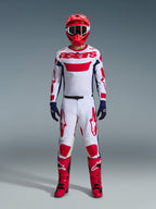 Piloto de motocross com o conjunto de equipamento Alpinestars 2026 Racer Riway, camisola branca com o logótipo "astars" a vermelho e painéis laterais azuis, calças brancas com grafismos vermelhos, capacete e botas de corrida vermelhos, luvas azul-escuras, equipamento de motocross completo e coordenado