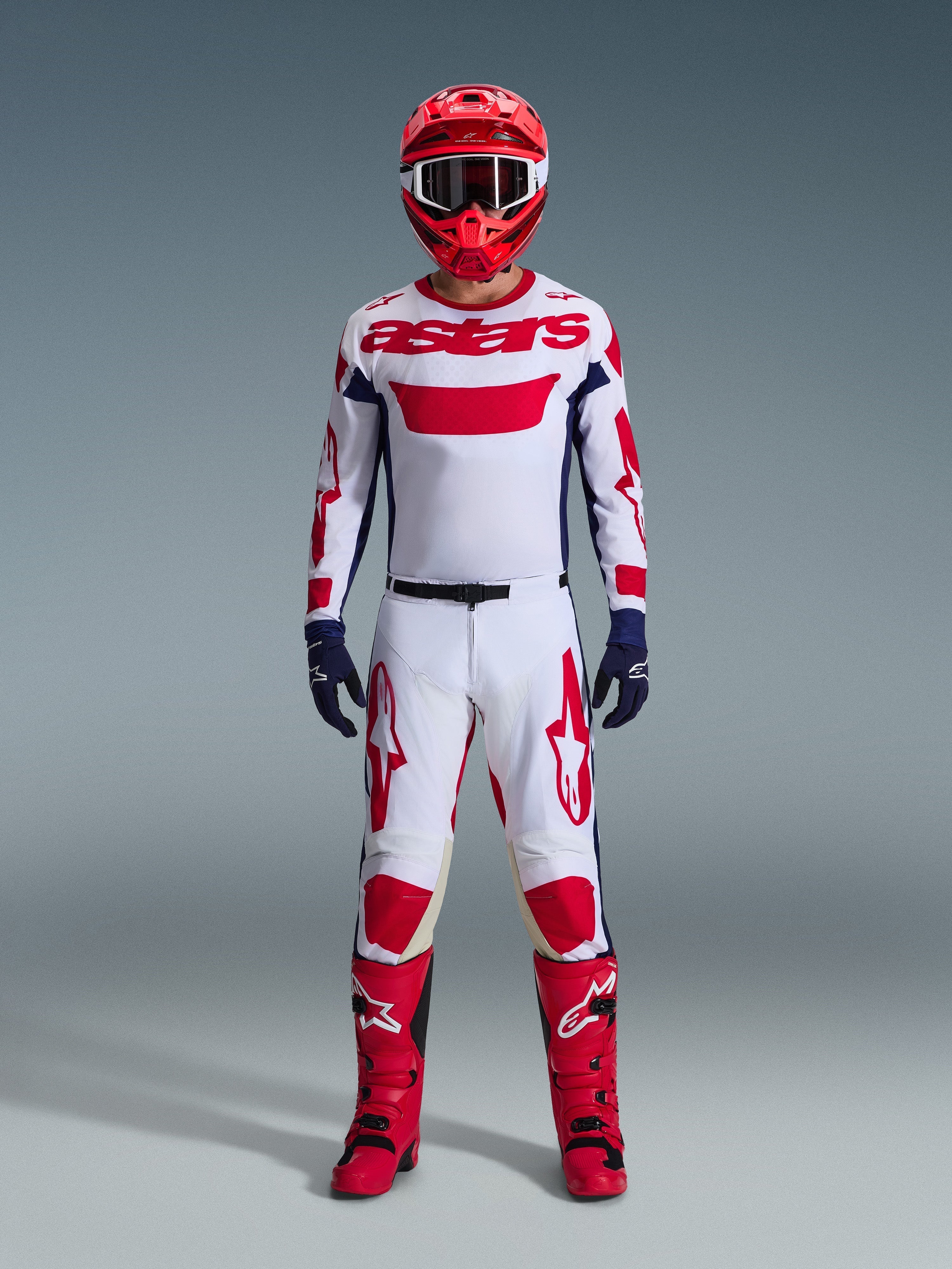 Piloto de motocross com o conjunto de equipamento Alpinestars 2026 Racer Riway, camisola branca com o logótipo "astars" a vermelho e painéis laterais azuis, calças brancas com grafismos vermelhos, capacete e botas de corrida vermelhos, luvas azul-escuras, equipamento de motocross completo e coordenado