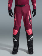 Alpinestars 2026 Racer Riway Pants, calças de motocross, vermelho coral escuro, usadas com luvas e botas pretas, com grandes logótipos Alpinestars em coral nas coxas, fivela de cintura preta e design de perna pré-curvada para corridas off-road