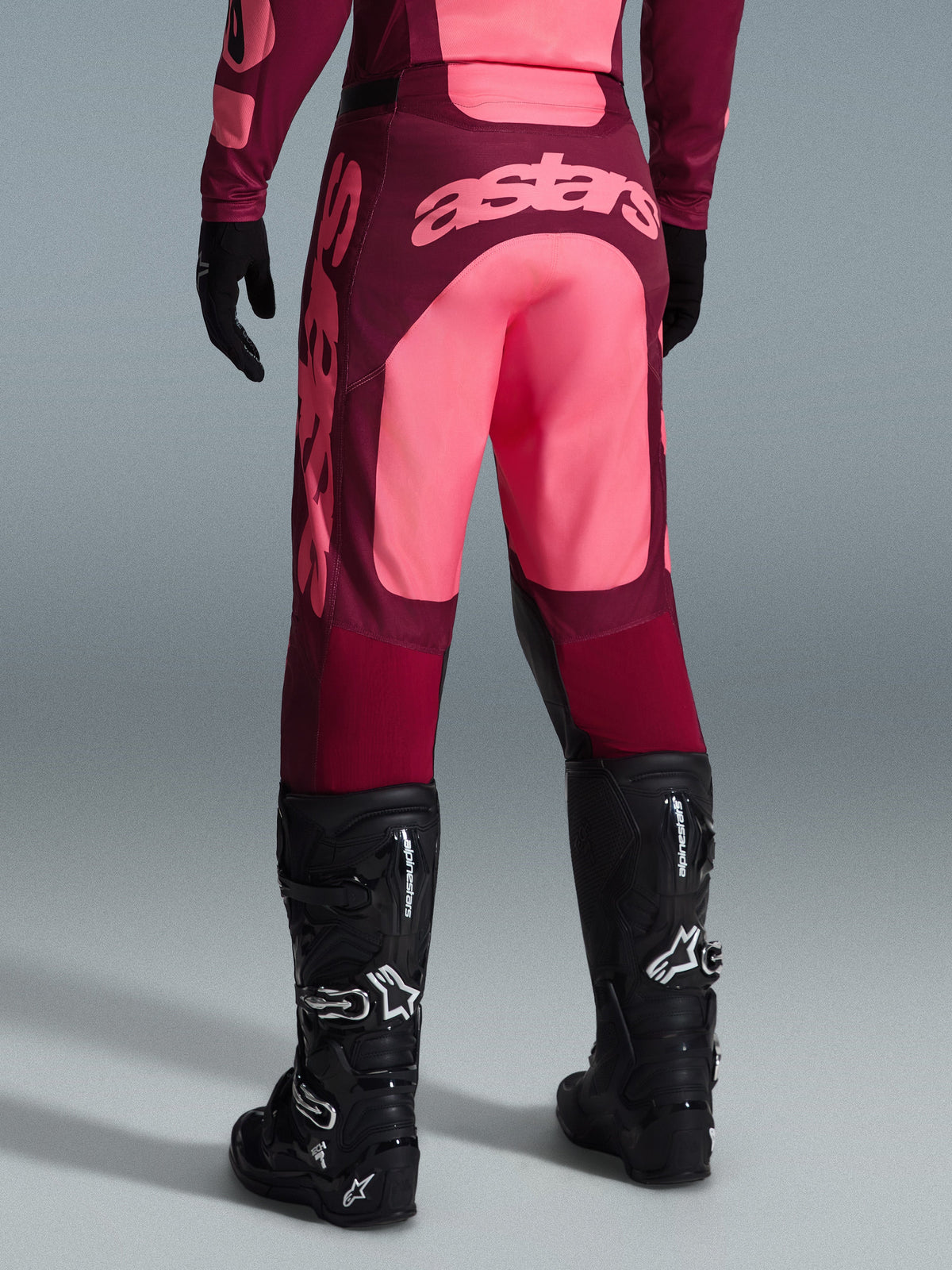 Alpinestars 2026 Racer Riway Pants, calças de motocross, vermelho escuro e coral, vista traseira com grande logótipo 'astars' em coral no assento, usadas com botas e luvas de motocross pretas, equipamento de performance para corridas todo-o-terreno