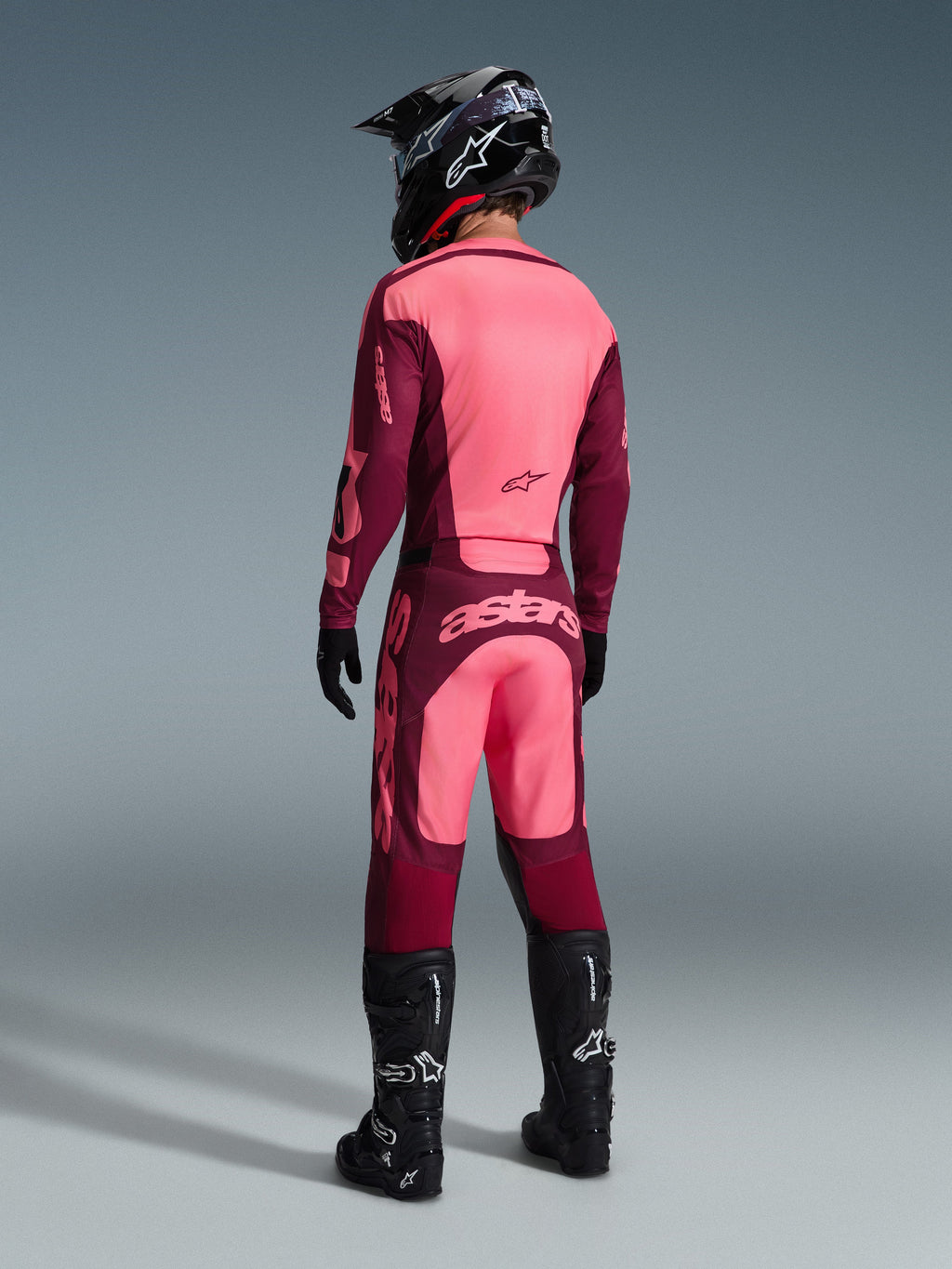 Piloto de motocross a usar o conjunto de equipamento Alpinestars 2026 Racer Riway visto de trás, em tons bordeaux e cor-de-rosa claro, camisola e calças com logótipos proeminentes, completo com capacete, luvas e botas pretas, fato de motocross coordenado