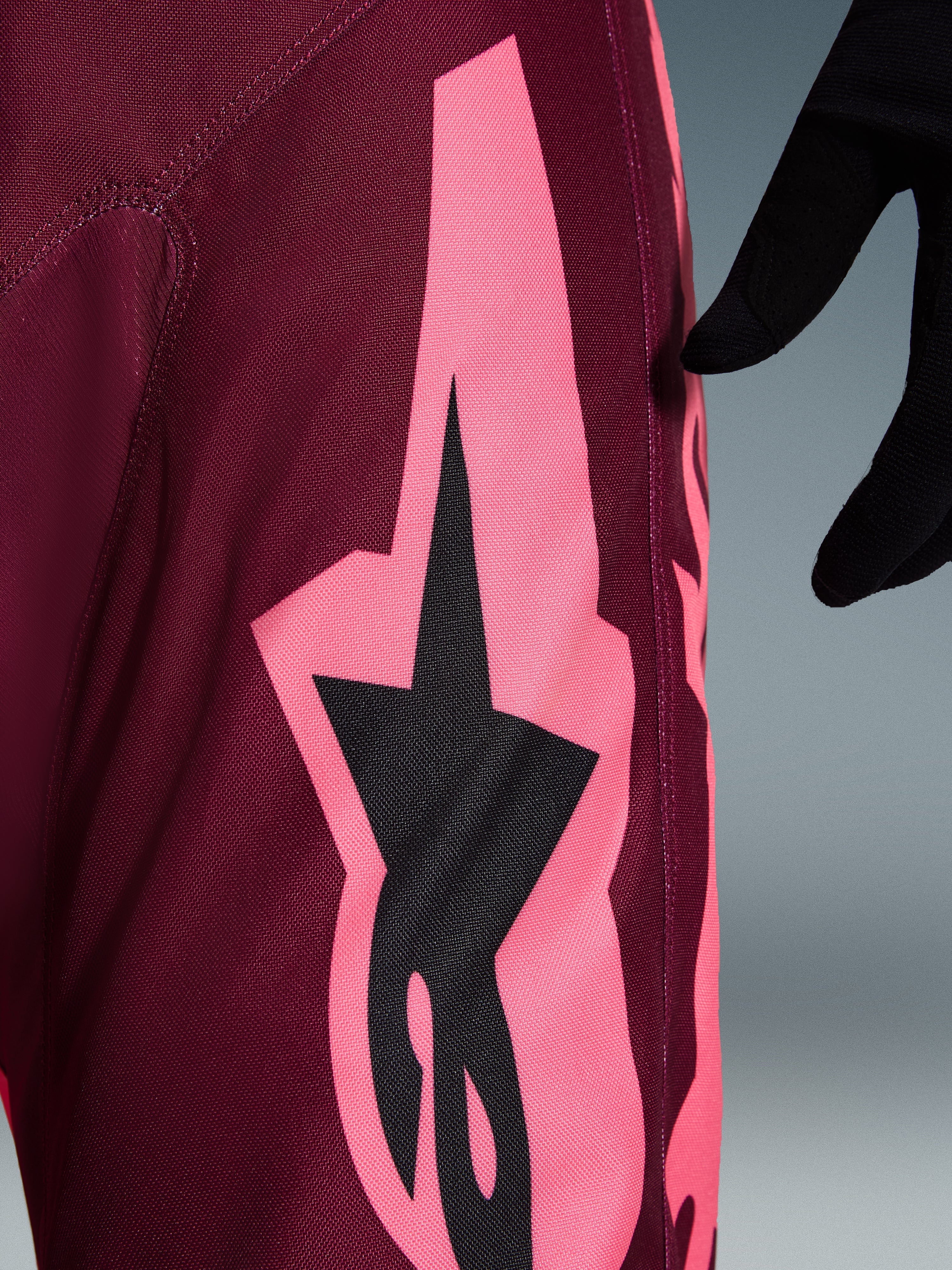 Calças Alpinestars 2026 Racer Riway, calças de motocross, Dark Red Coral, vista de pormenor com um grande gráfico angular rosa coral e logótipo de estrela preto, destacando a textura do tecido durável e as costuras de performance