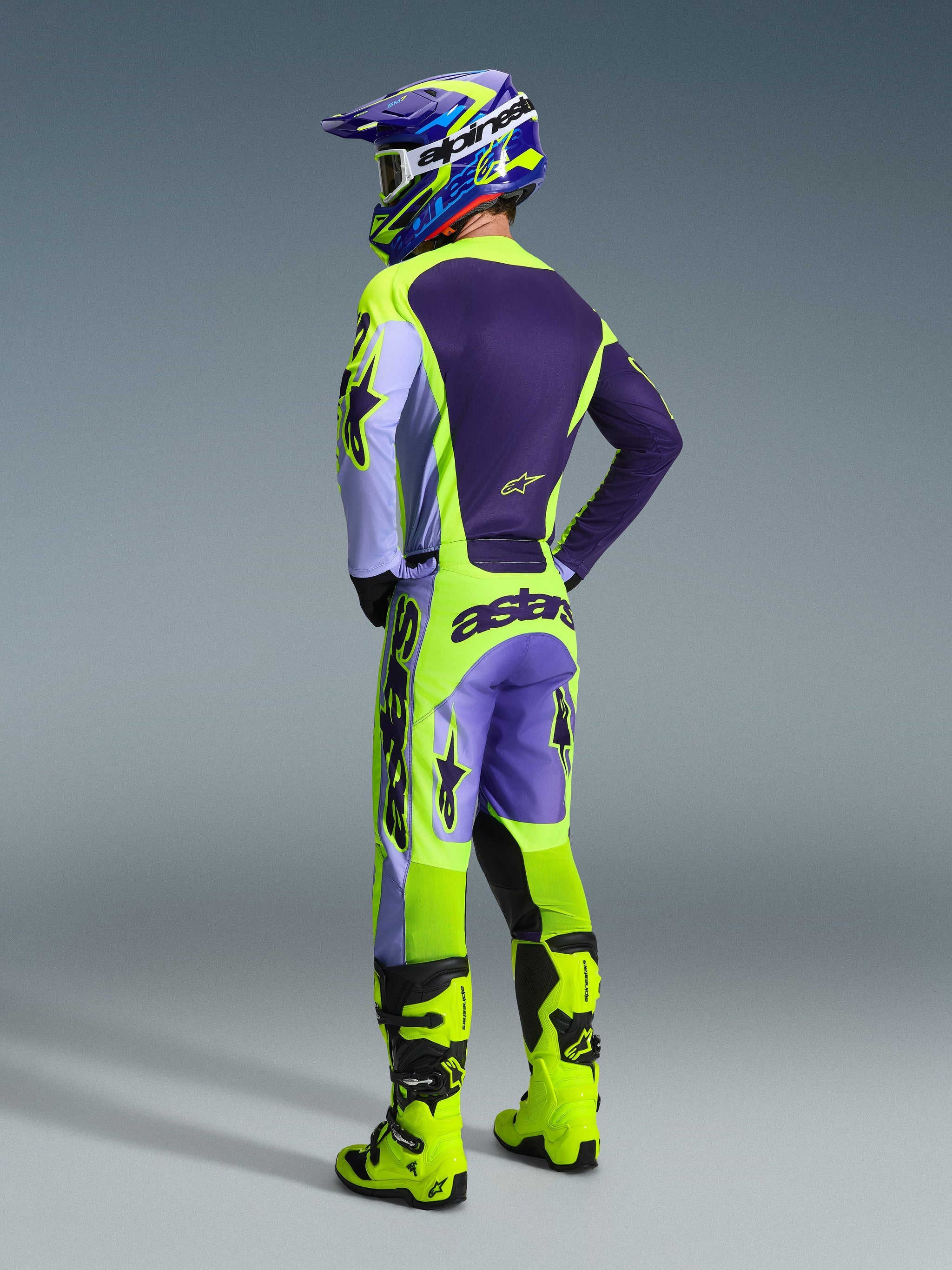 Motocross rider from behind wearing the Alpinestars 2026 Racer Portl Pants in Purple Yellow Fluo, styled with a matching purple and yellow jersey, helmet, and bright yellow racing boots, featuring bold 'astars' branding on the seat and star logos on the legs.