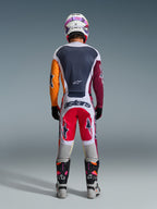Piloto de motocross visto de costas a usar as calças Alpinestars Racer Portl 2026 em vermelho, cinzento claro e cinzento escuro, com a marca "astars" no assento, combinadas com uma camisola multicolorida, capacete branco e botas de competição técnicas.