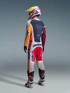 Piloto de motocross visto de costas a usar equipamento de corrida completo, calças Alpinestars 2026 Racer Portl em Vermelho, Cinzento Claro e Cinzento Escuro, camisola a condizer com mangas cor de laranja e bordeaux, capacete com grafismos multicoloridos, botas de motocross cinzentas e pretas com detalhes cor-de-rosa, conjunto de motocross profissional