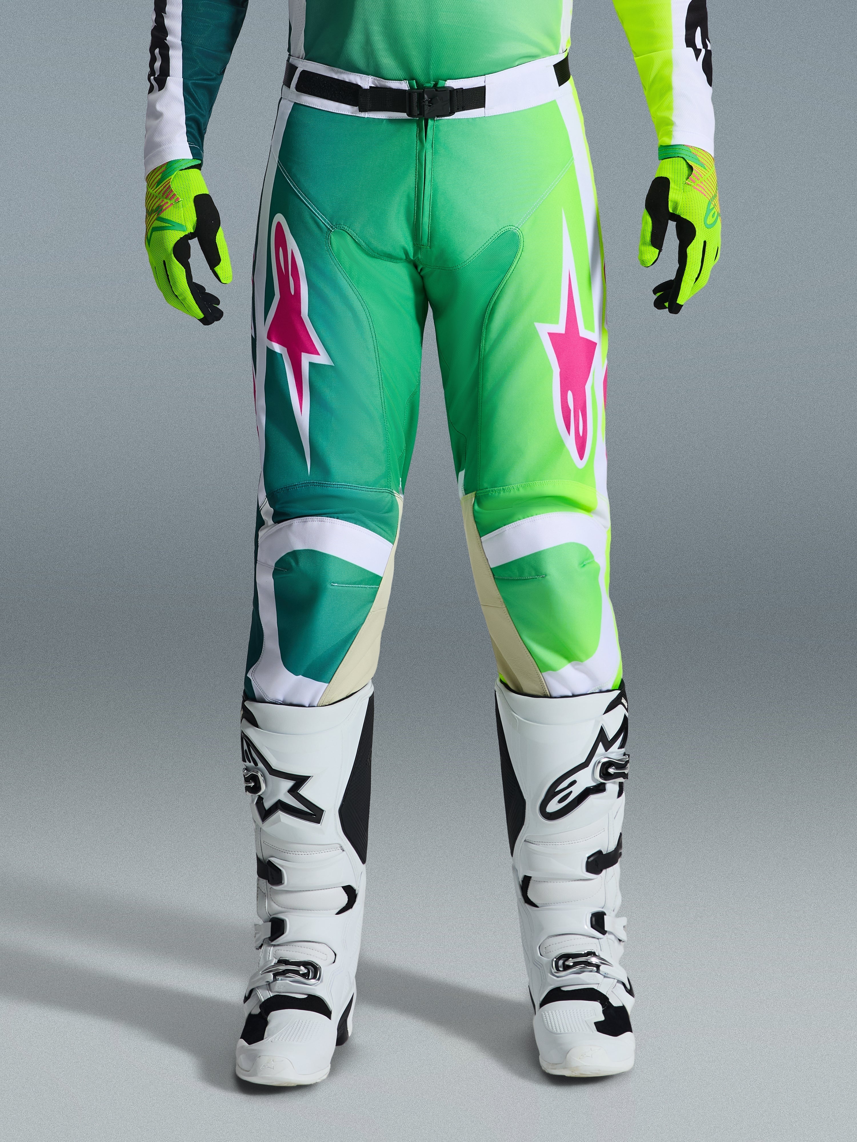 Alpinestars 2026 Racer Portl Pants, calças de motocross, verdes, brancas e pretas com logótipos de estrela cor-de-rosa, usadas com botas brancas e pretas e luvas verde-lima, com um design em degradê e ajuste ergonómico para corridas de off-road