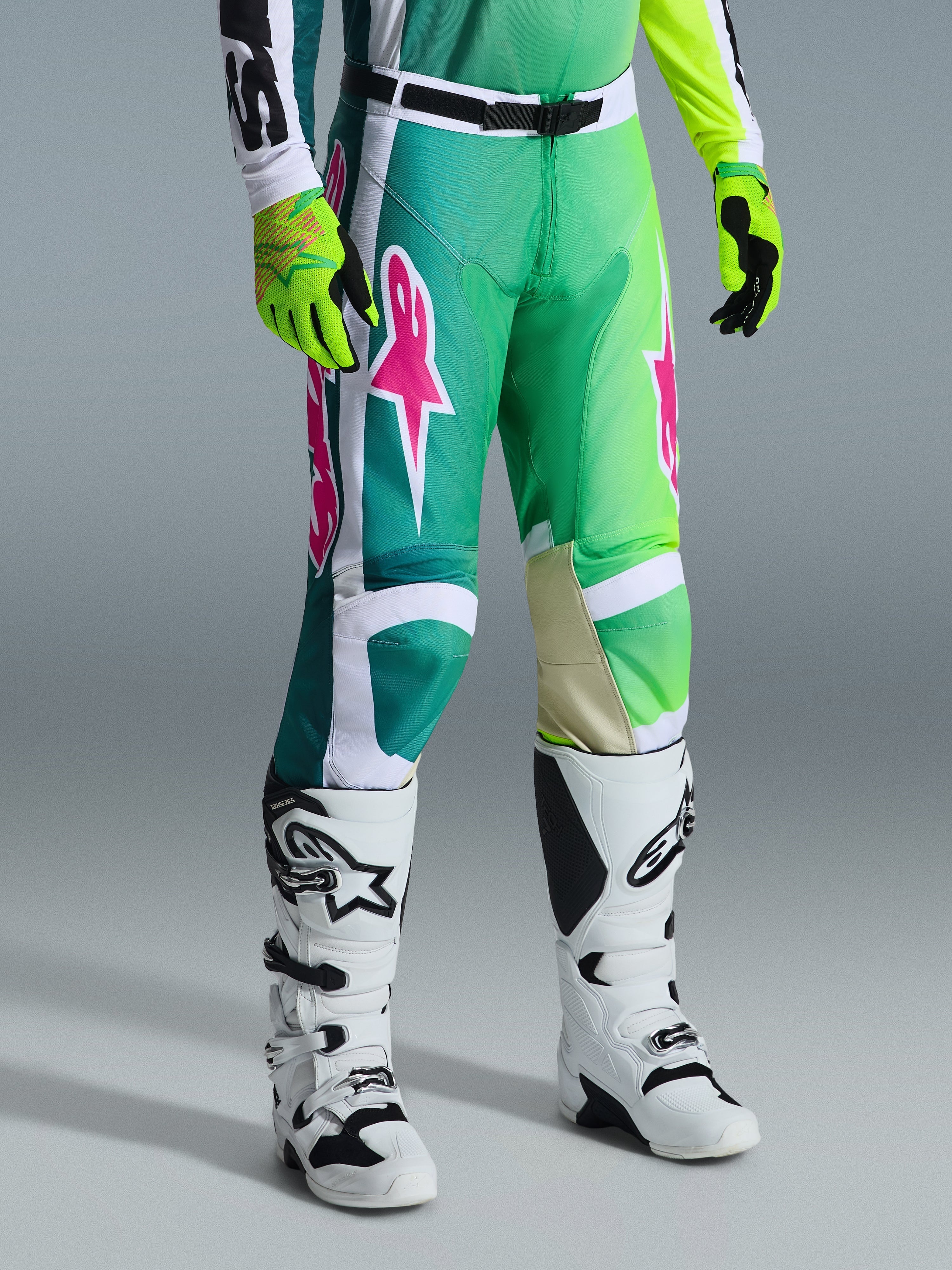 Alpinestars 2026 Racer Portl Pants, calças de motocross, verdes, brancas e pretas com logótipos cor-de-rosa, usadas com botas brancas e luvas amarelo néon, com padrões gráficos arrojados e uma fivela de cintura preta ajustável, equipamento de performance para condução todo-o-terreno