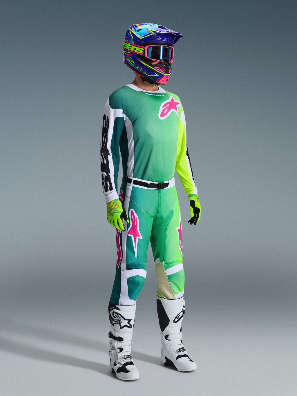 Piloto de motocross a usar calças Alpinestars 2026 Racer Portl e camisola a condizer em verde, branco e preto com detalhes do logótipo em rosa, capacete colorido, botas de competição brancas e pretas e luvas verde néon, conjunto de equipamento completo para corridas off-road