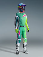Piloto de motocross a usar calças Alpinestars 2026 Racer Portl e camisola a condizer em verde, branco e preto com detalhes do logótipo em rosa, capacete colorido, botas de competição brancas e pretas e luvas verde néon, conjunto de equipamento completo para corridas off-road