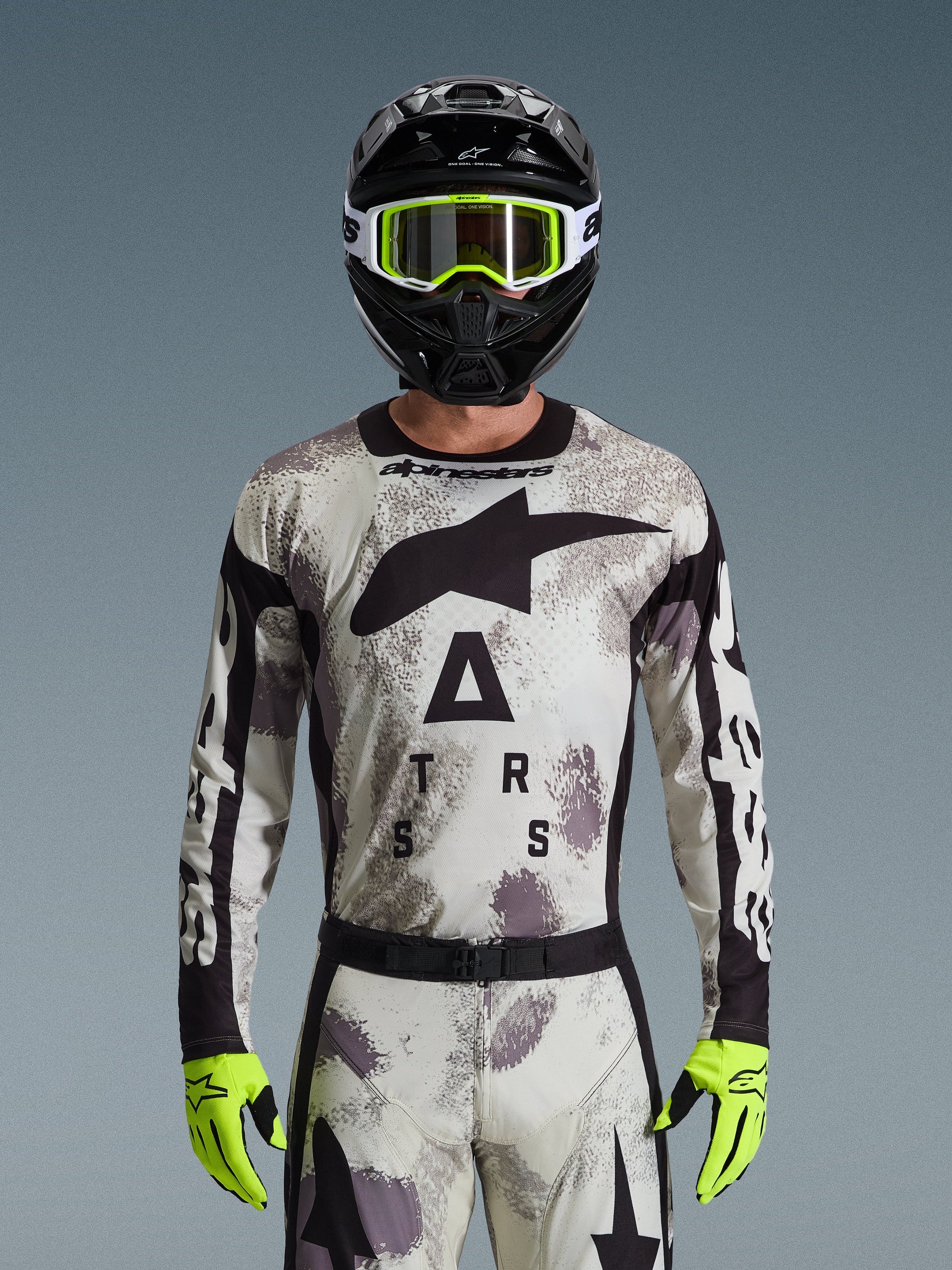 Piloto de motocross a usar a Camisola Alpinestars Racer Lahnd 2026, padrão Sand Camo em cinzento e branco, grande logótipo Astar preto e branding no peito, combinado com capacete preto, óculos e luvas amarelo néon, equipamento de performance para condução off-road